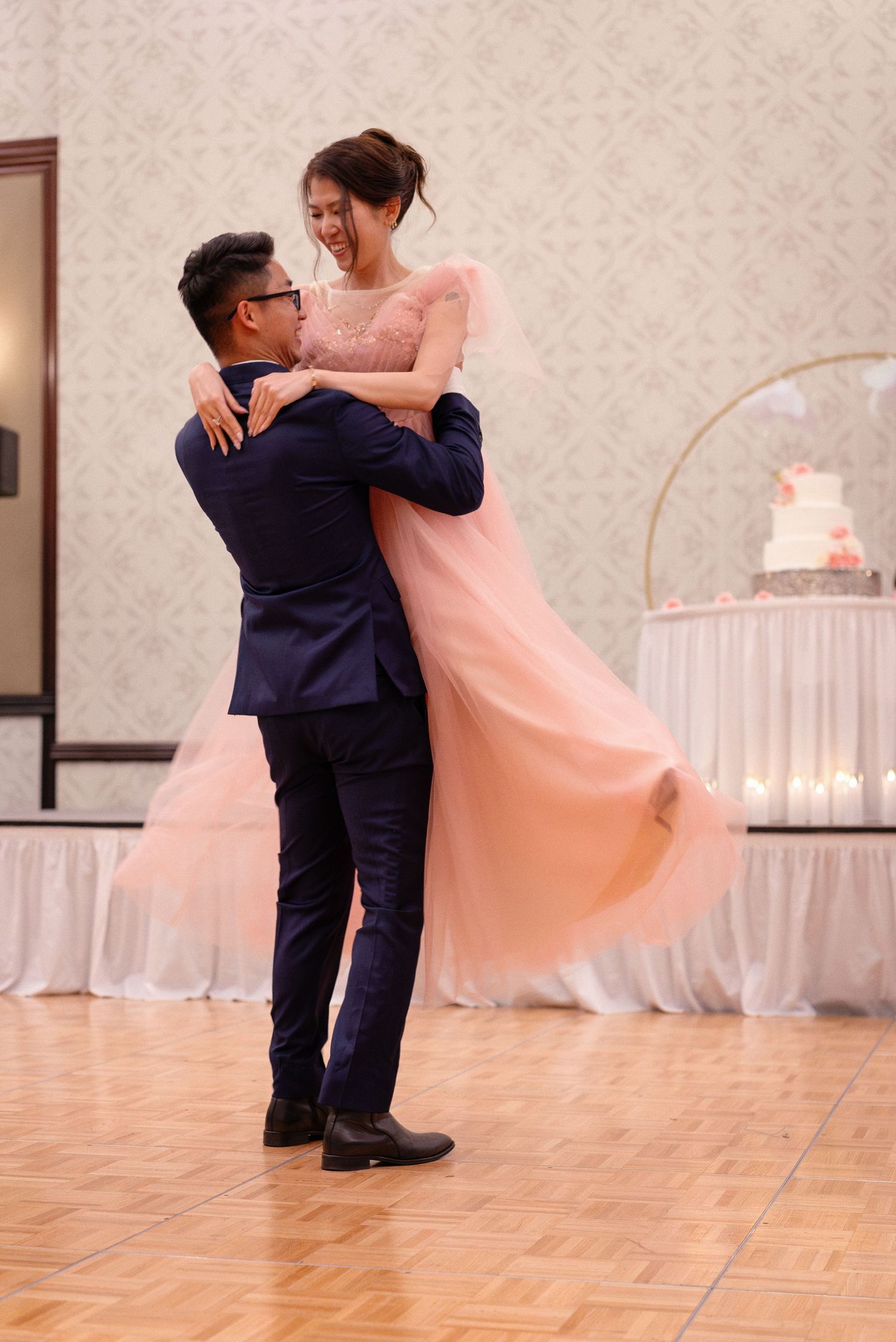 Groom lifts bride in a pink flowing gown during their first dance at a wedding reception.