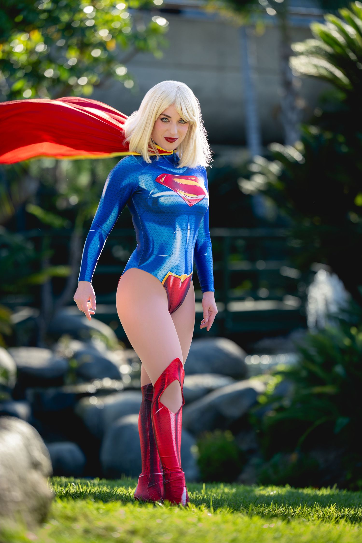 Woman in Supergirl cosplay costume with red cape and blue bodysuit posing outdoors in a garden setting.