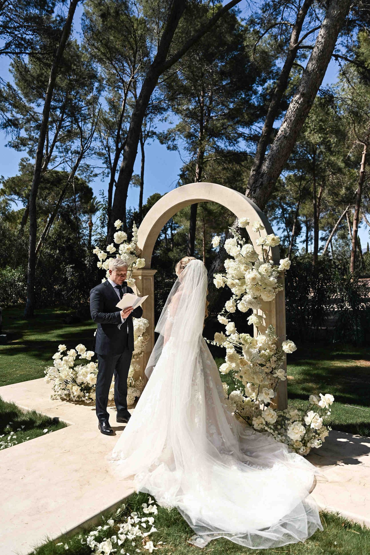 Groom reading personalized vows to his bride under a beautiful white floral arch at a destination wedding ceremony in Spain.