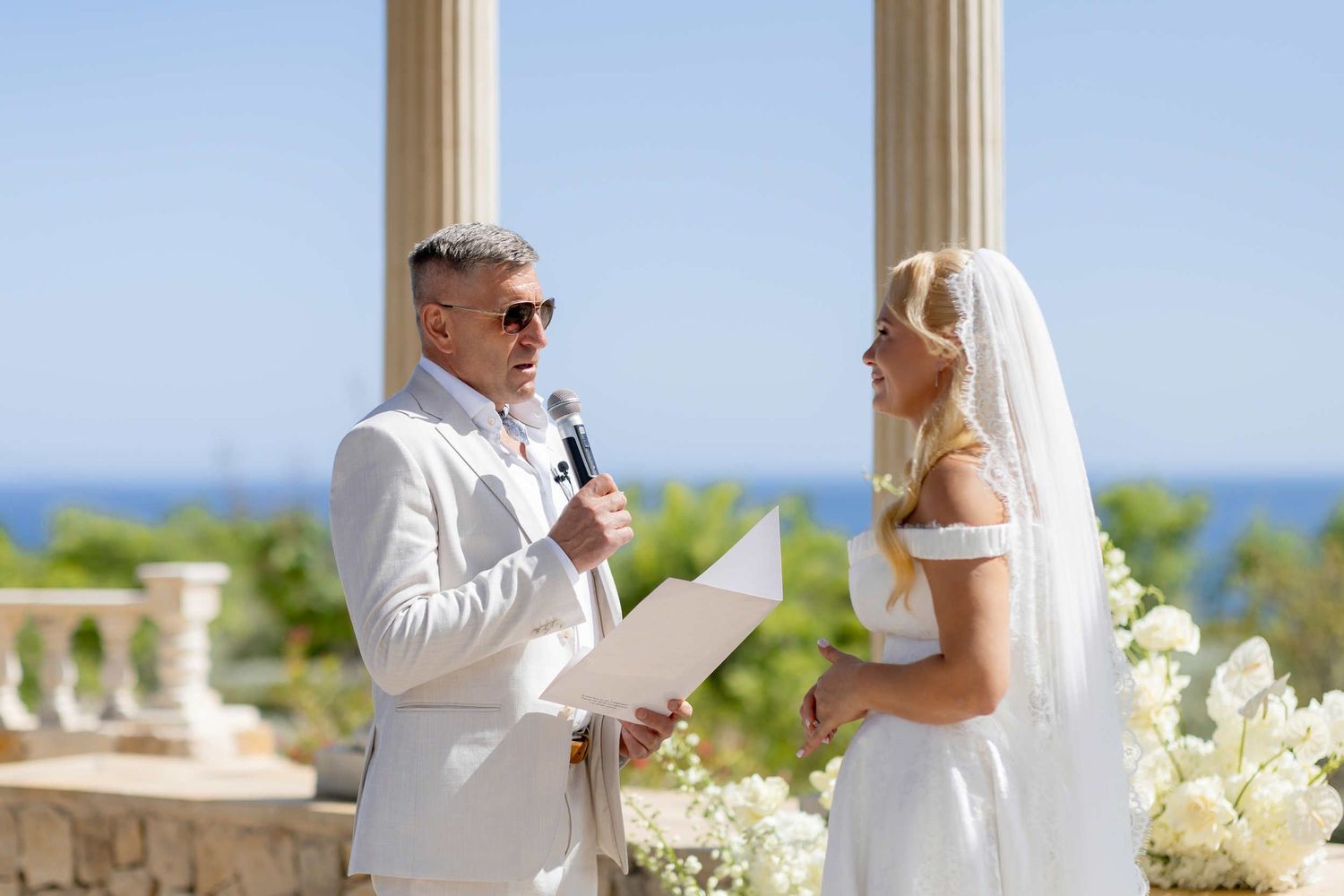 Groom in sunglasses reading vows into a microphone to his smiling bride during a sunny coastal ceremony on the Costa Blanca.