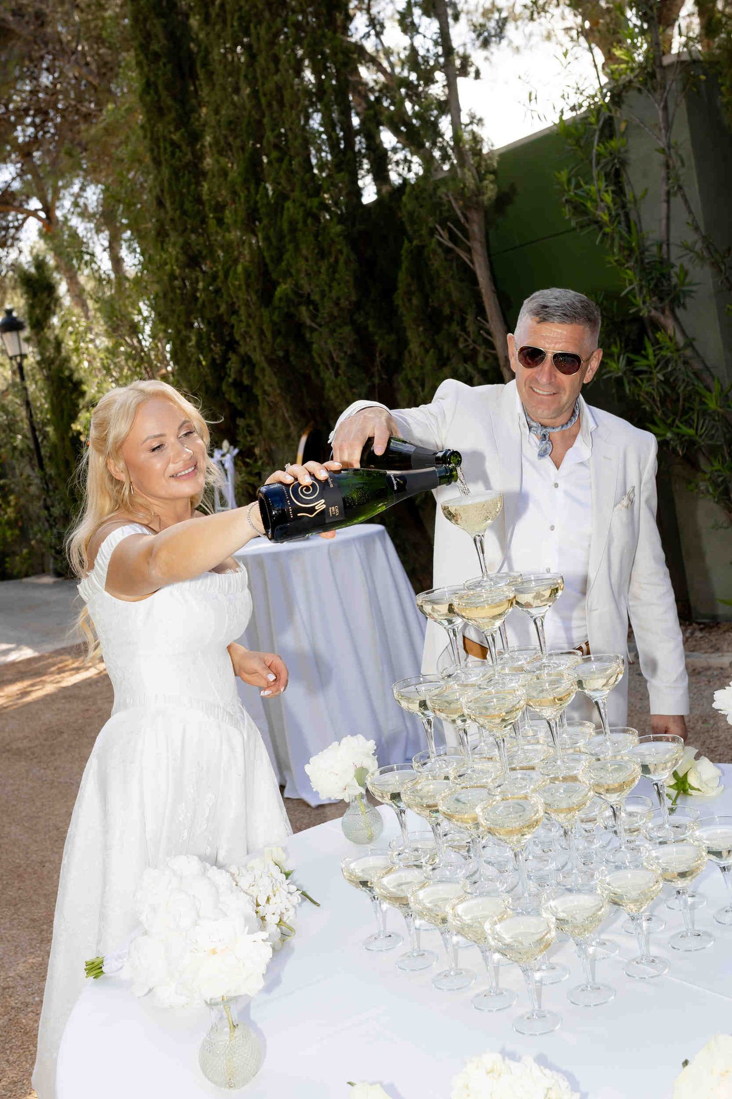 Bride and groom pouring champagne into a tall glass tower during a sunny outdoor wedding reception at Finca Marques de Montemolar