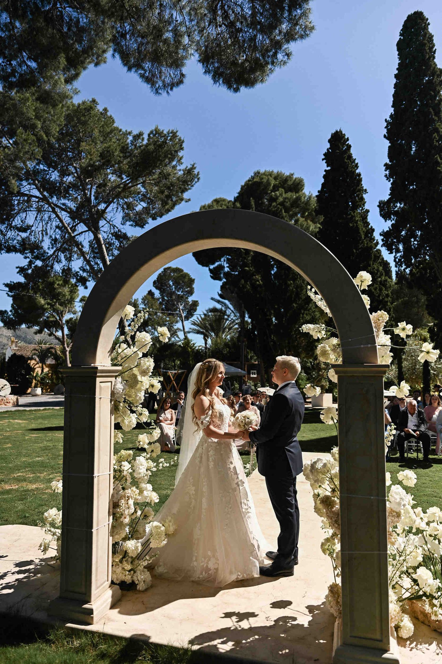 Bride and groom exchanging vows under a stone arch with wedding guests seated outdoors at Finca El Senyoret