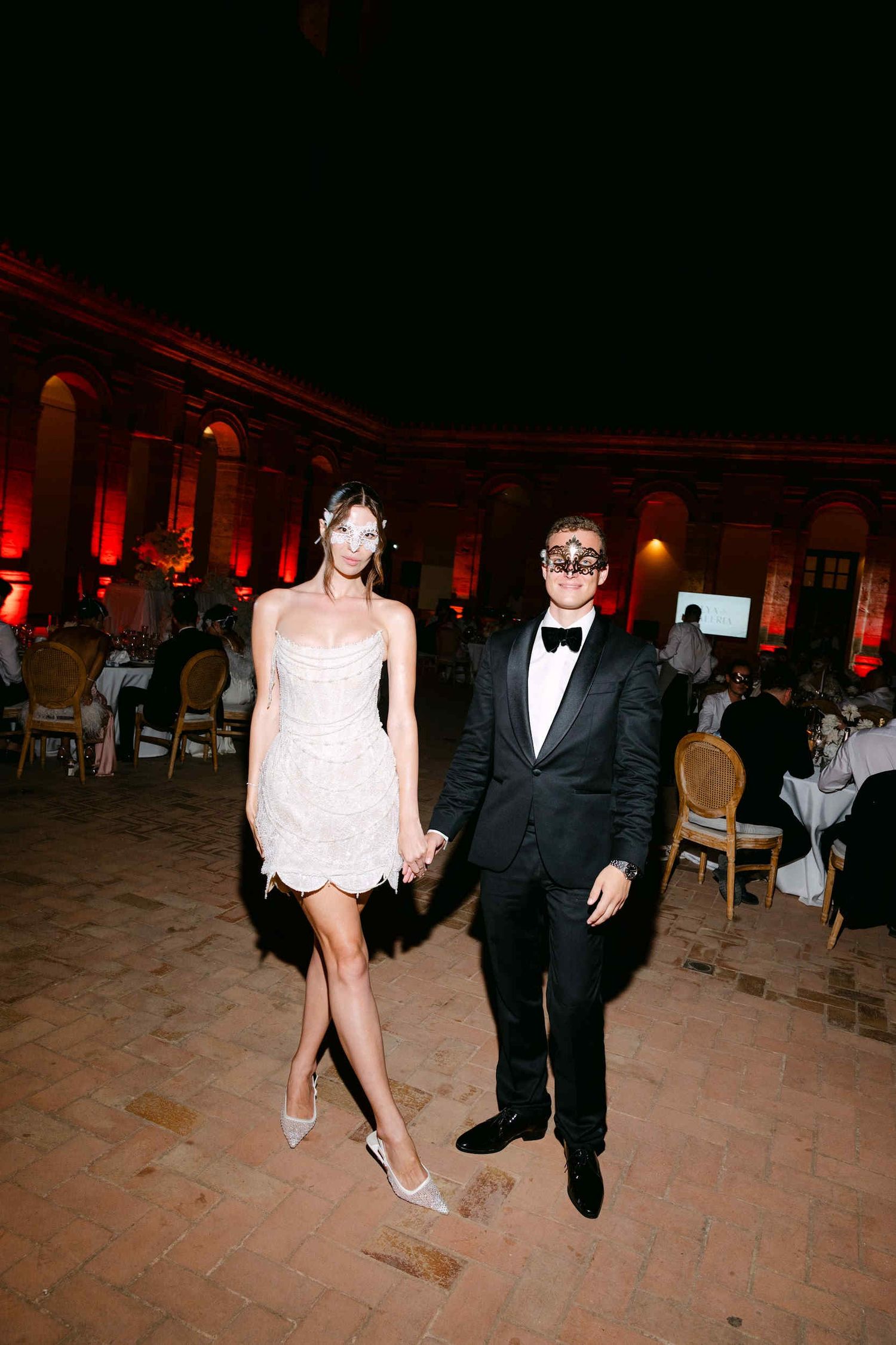 Bride in a short dress and groom in a black tuxedo wearing Venetian masks at their La Cartuja wedding party.