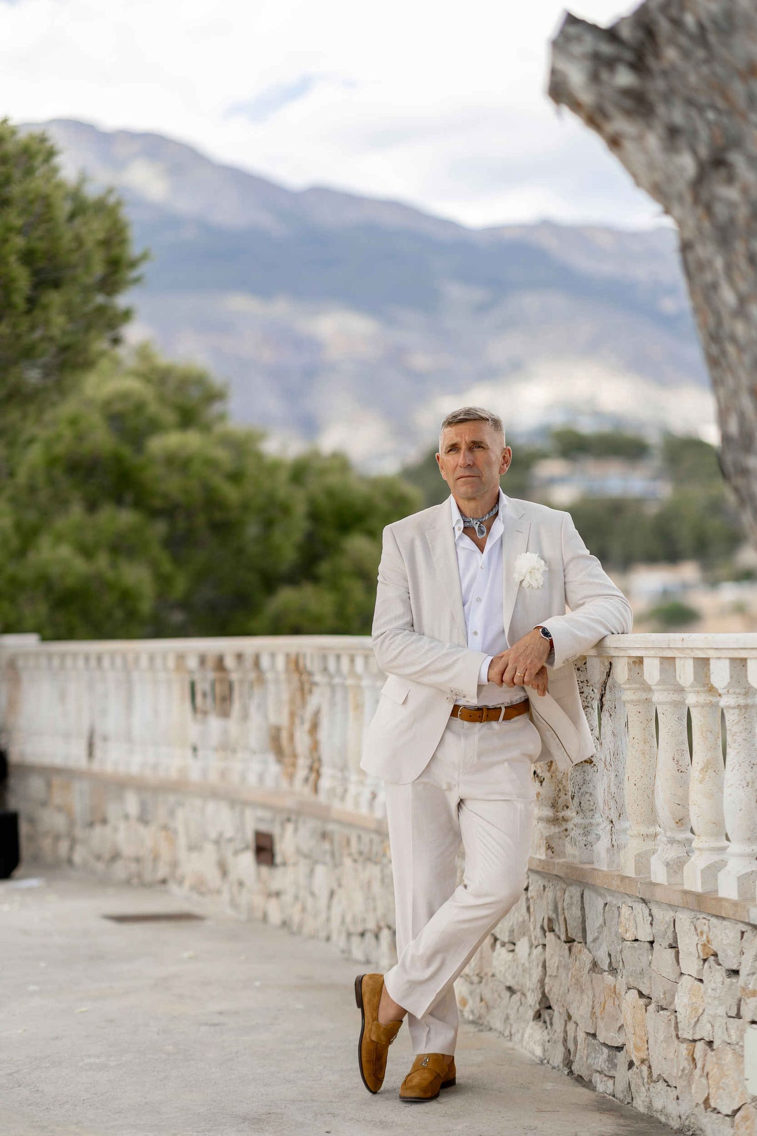 Groom in a stylish white suit leaning against a stone railing with mountain views during his destination wedding in Altea.