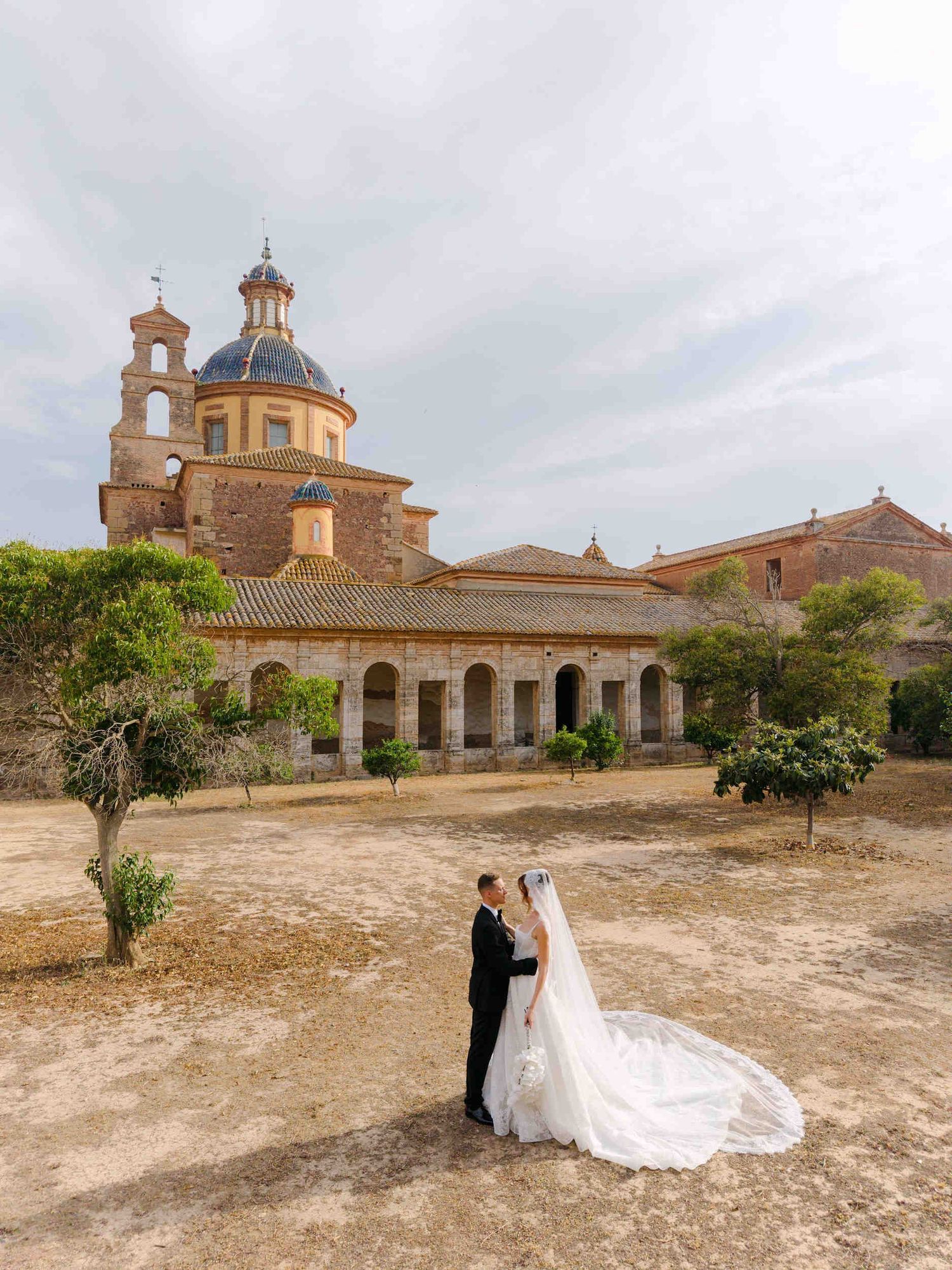 Newlyweds posing on a sandy courtyard with the grand dome and historic stone architecture of a Spanish monastery behind them.