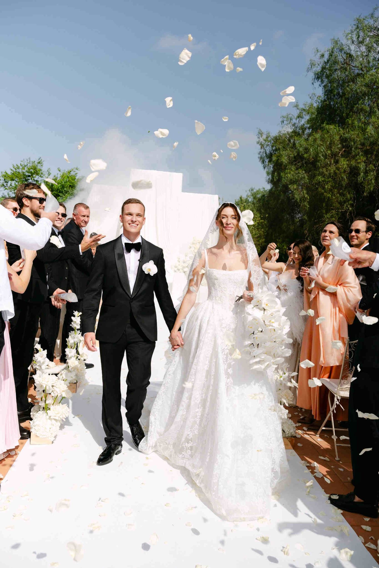 Smiling newlyweds holding hands as guests toss white rose petals during their sunny outdoor wedding exit in monastery cartuja ara christi