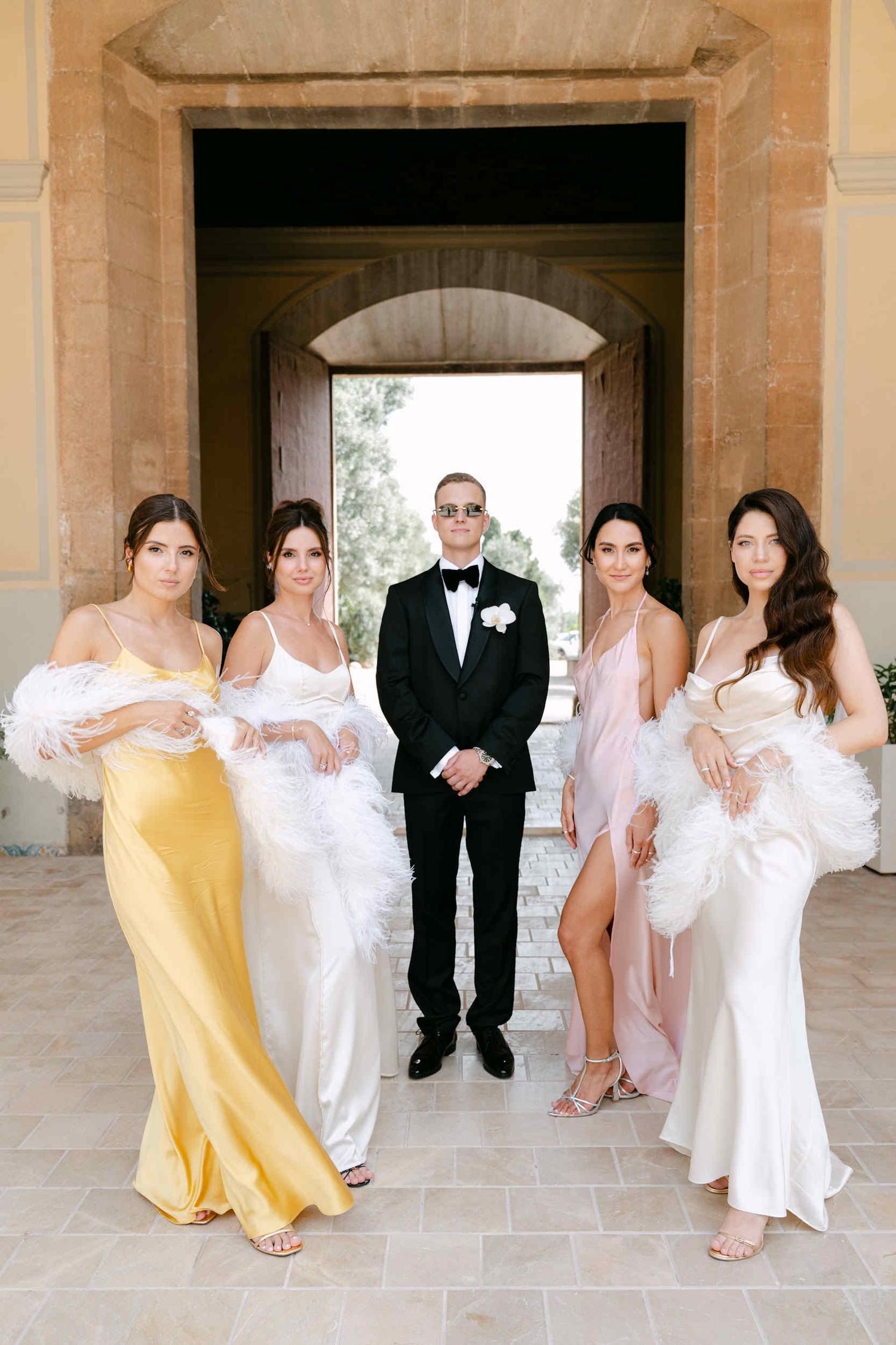 Groom wearing sunglasses standing with bridesmaids in pastel dresses and feather boas inside a La Cartuja stone archway.