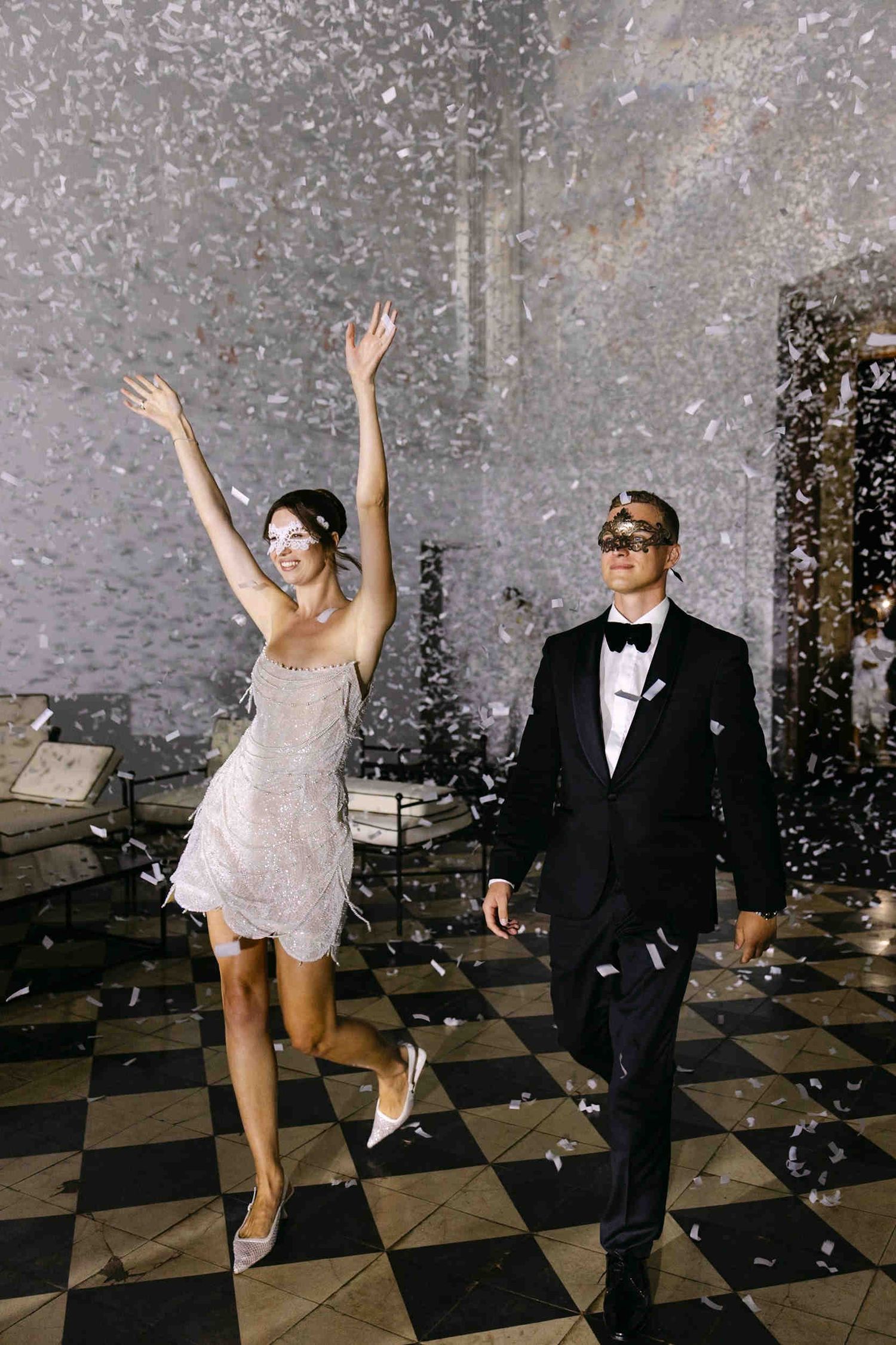 Newlyweds wearing masquerade masks dancing and celebrating under falling white confetti on a checkered floor at La Cartuja.