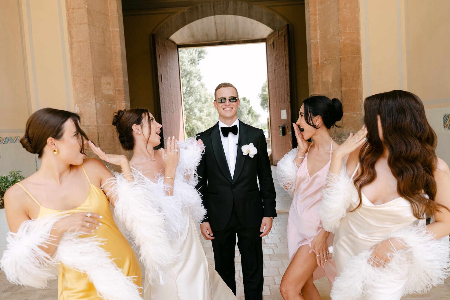 Groom in a black tuxedo laughing with bridesmaids in silk dresses and feather boas under a stone archway in Valencia.