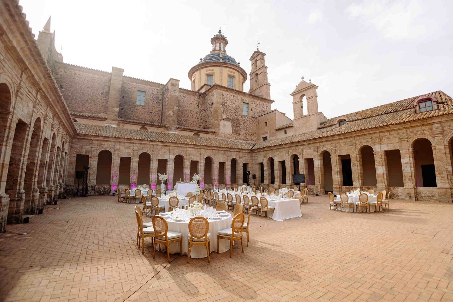 Outdoor wedding reception in a grand stone courtyard with round tables, wooden chairs, and a historic domed roof in Valencia.