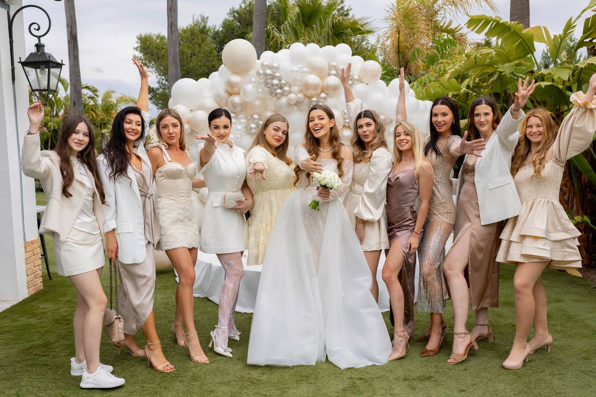 A bride and her bridesmaids pose playfully reaching toward the camera against a white balloon backdrop at an Alicante estate.