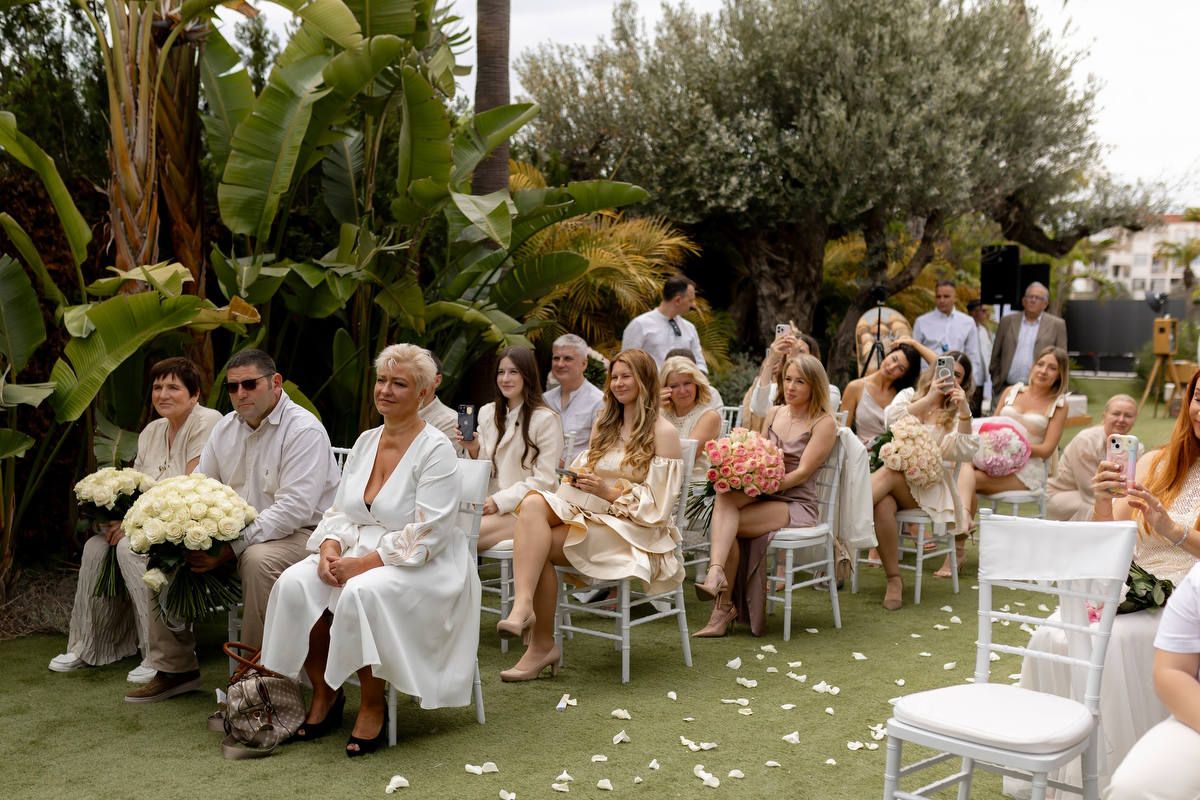 Seated wedding guests holding lush rose bouquets smile during an outdoor garden ceremony at an Alicante country estate.