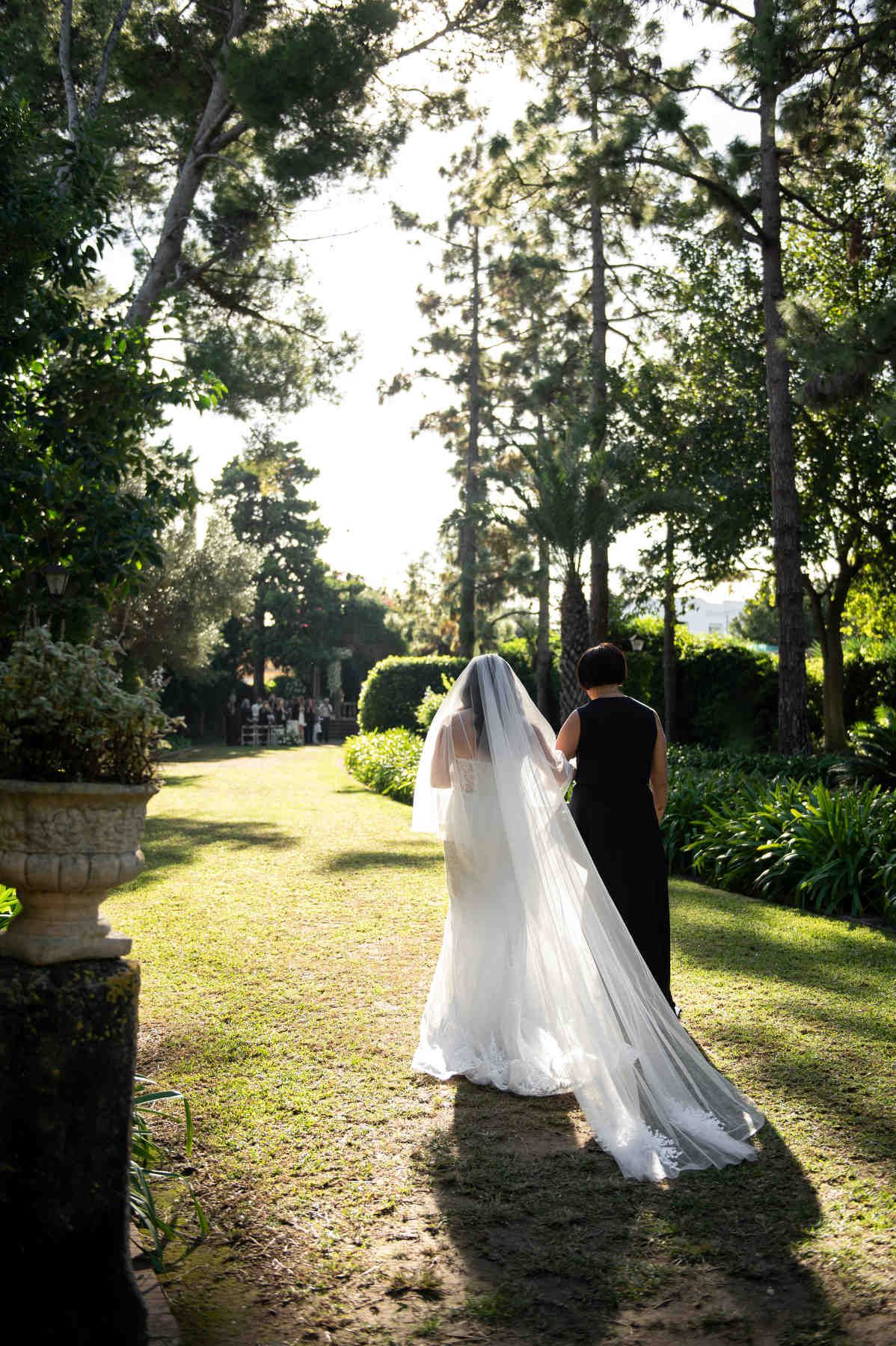 Bride walking down the garden aisle with her mother and long veil under pine trees at Casa Santonja on the Costa Blanca