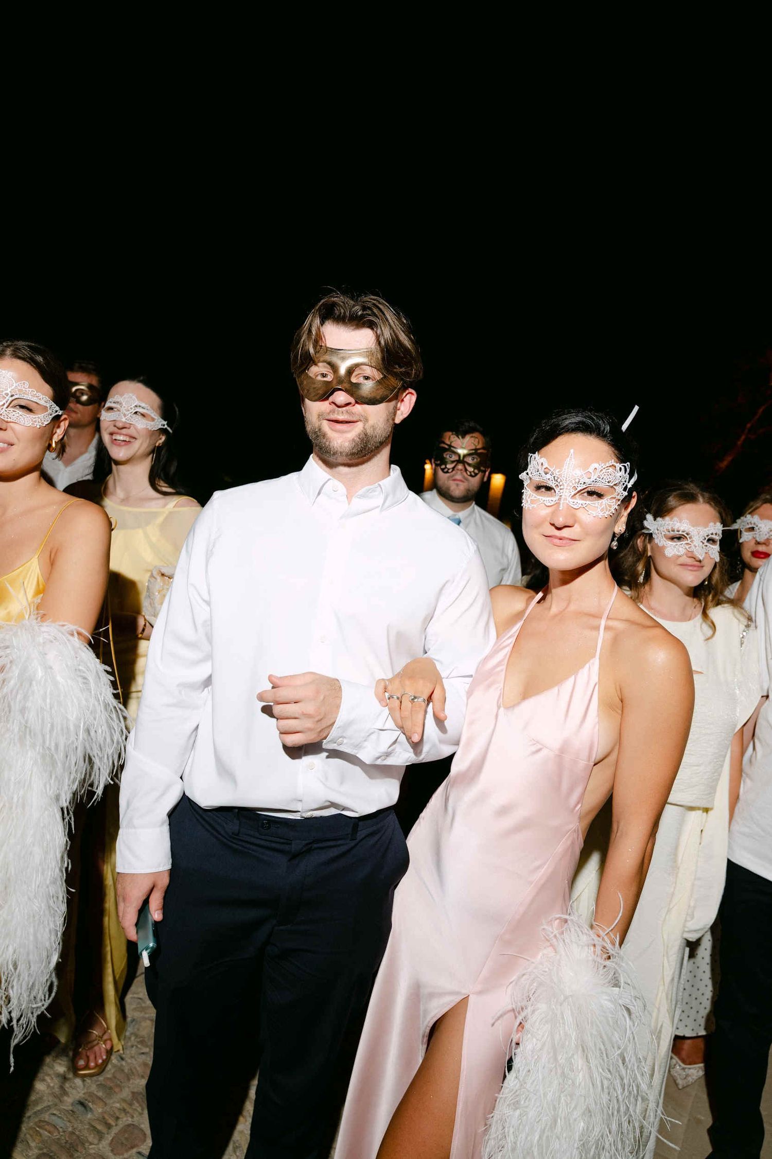 A male and female wedding guest wearing gold and white lace masquerade masks smiling at the La Cartuja evening reception.