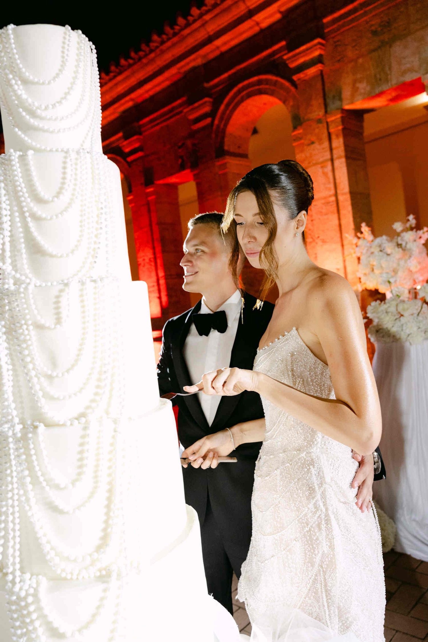 Close-up of the couple cutting their pearl-decorated wedding cake at La Cartuja with warm red architectural lighting.
