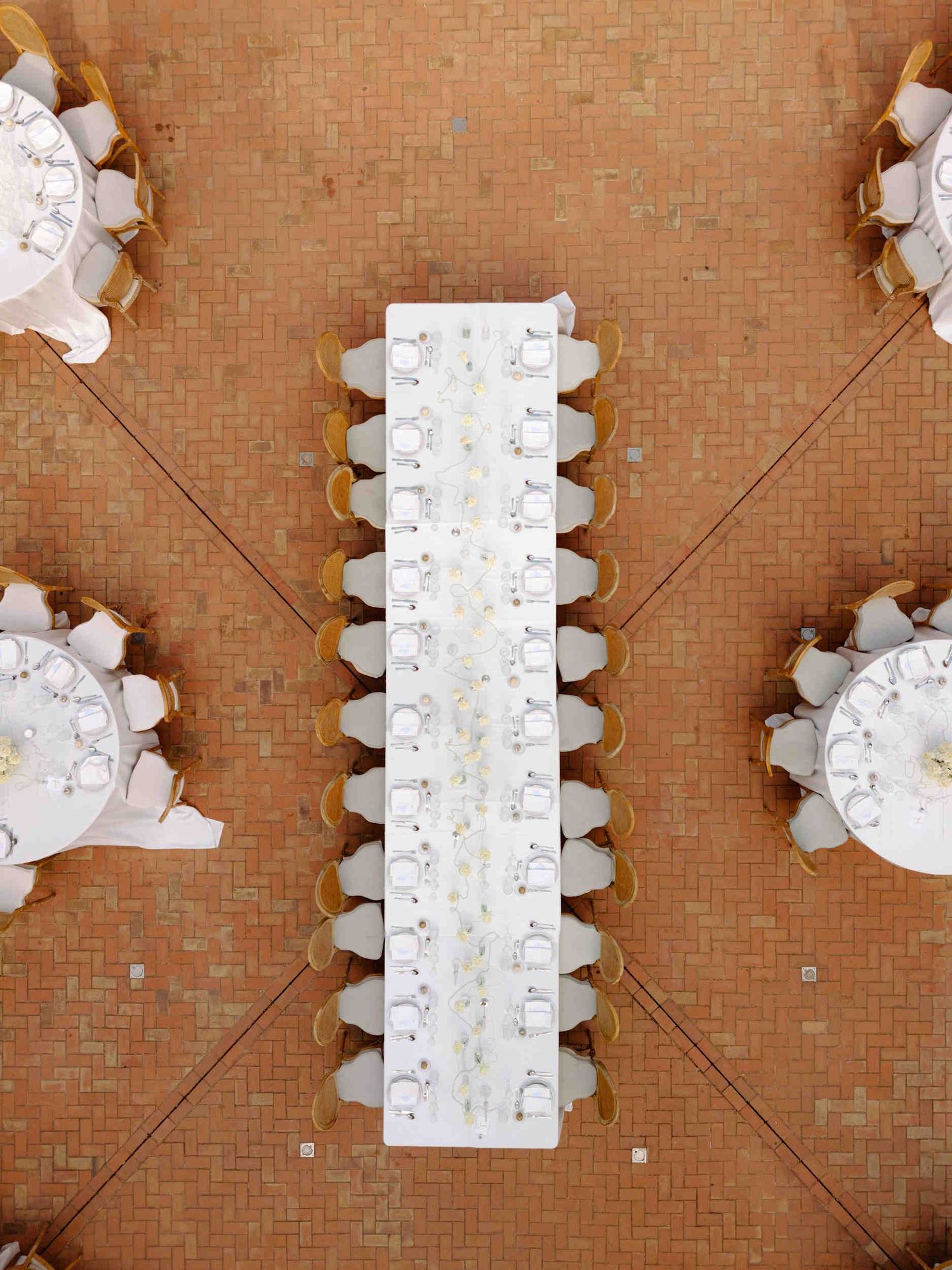 Top-down view of a long white wedding dinner table surrounded by wooden chairs on a terracotta tiled courtyard floor in Spain.