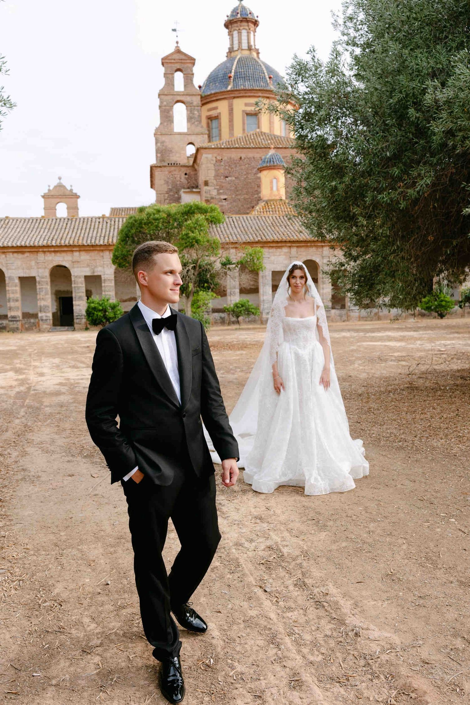 Groom in a sharp black tuxedo stands in the foreground while his bride poses softly in the background at the historic estate.