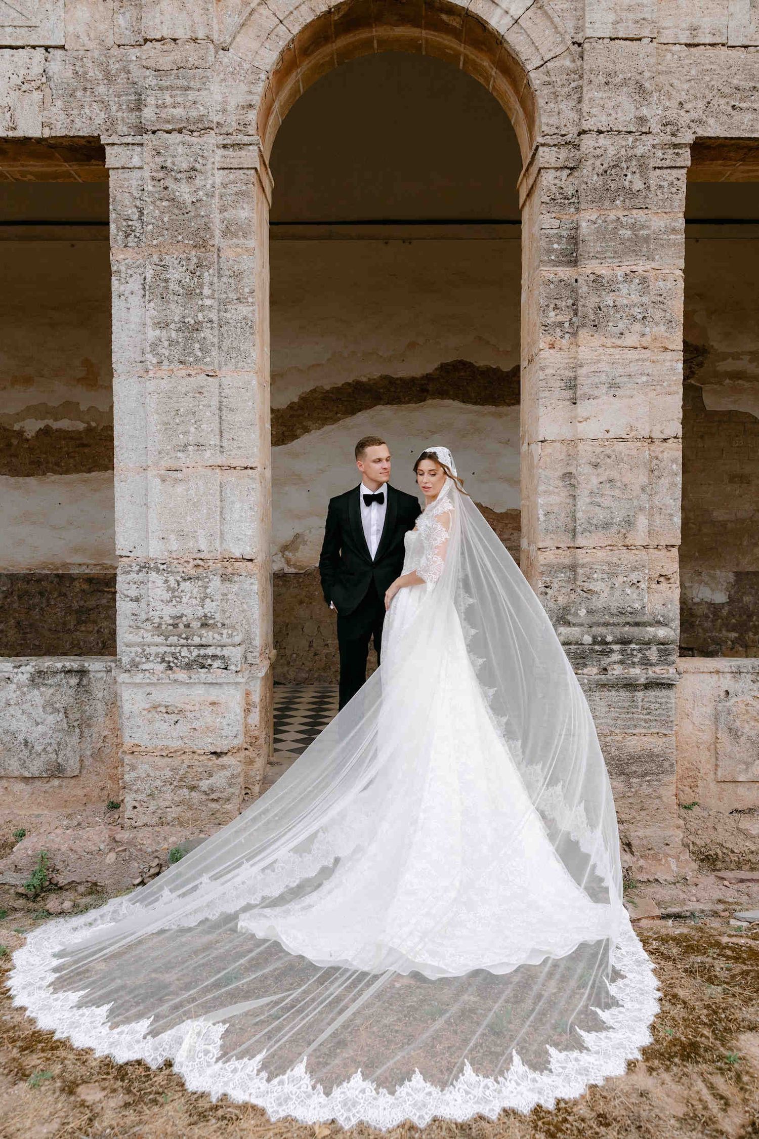 Bride wearing a long flowing veil and groom in a tuxedo posing inside a rustic stone archway at a historic cartuja monastery
