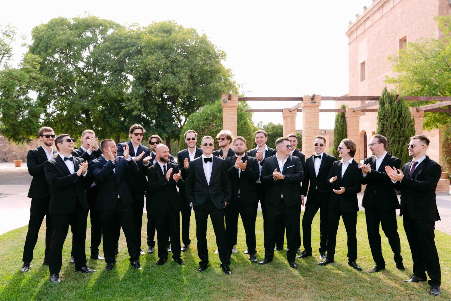 Groomsmen in black suits clapping for the groom on a green lawn outside the La Cartuja de Ara Christi estate.