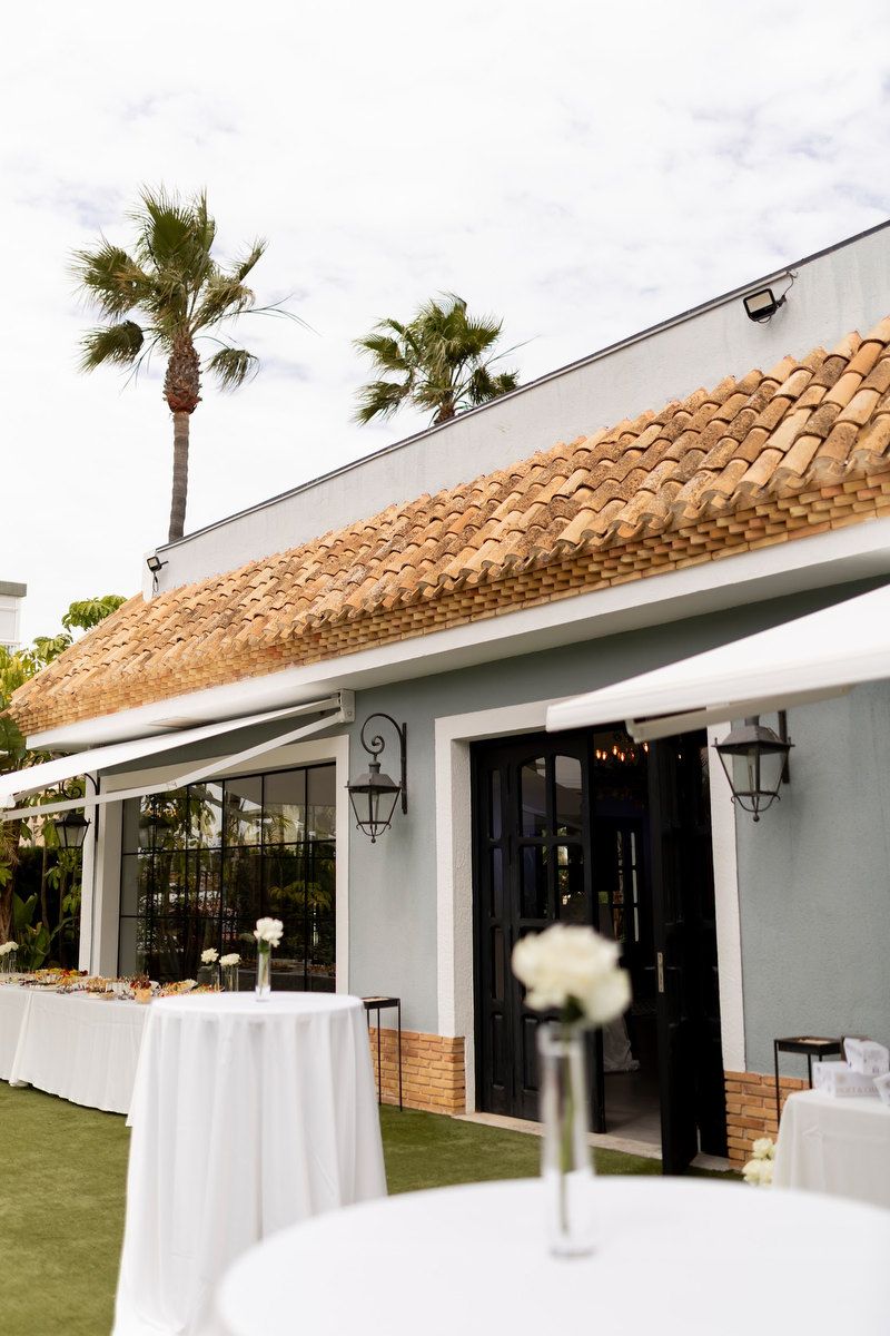High cocktail tables with single white roses stand on a green lawn outside a historic light blue finca in Alicante.