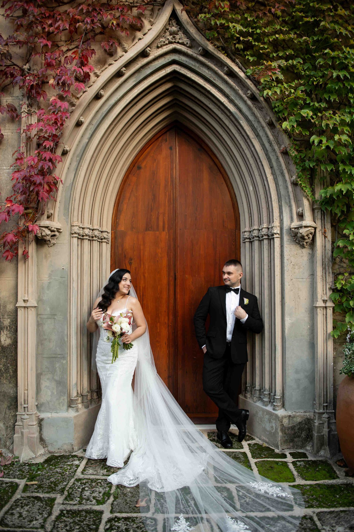 Bride and groom posing by large wooden doors in an ivy-covered stone archway, styled by a Casa Santonja wedding planner.