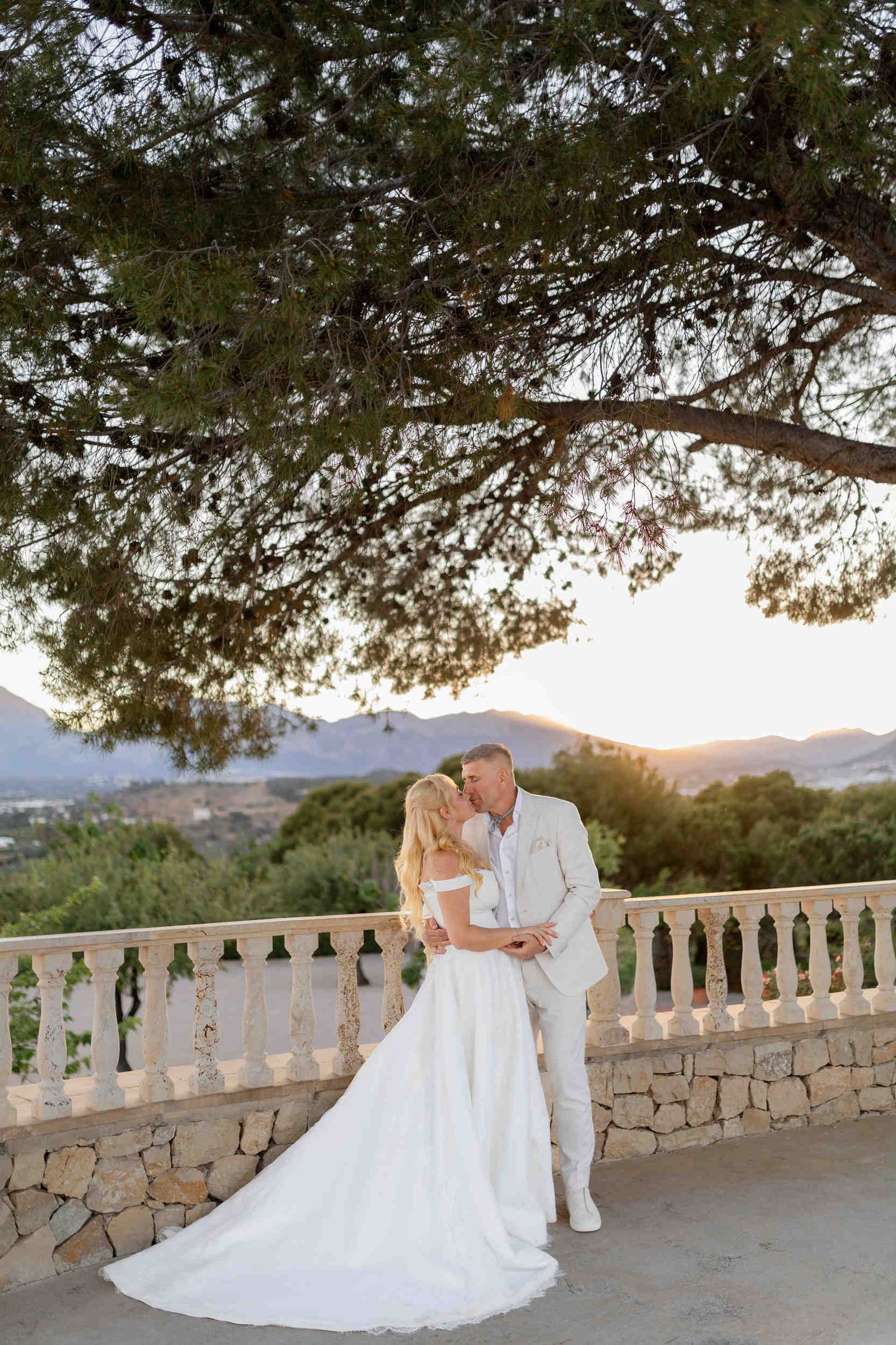 Bride and groom kissing under a pine tree on a stone terrace at sunset during their beautiful wedding in Altea.