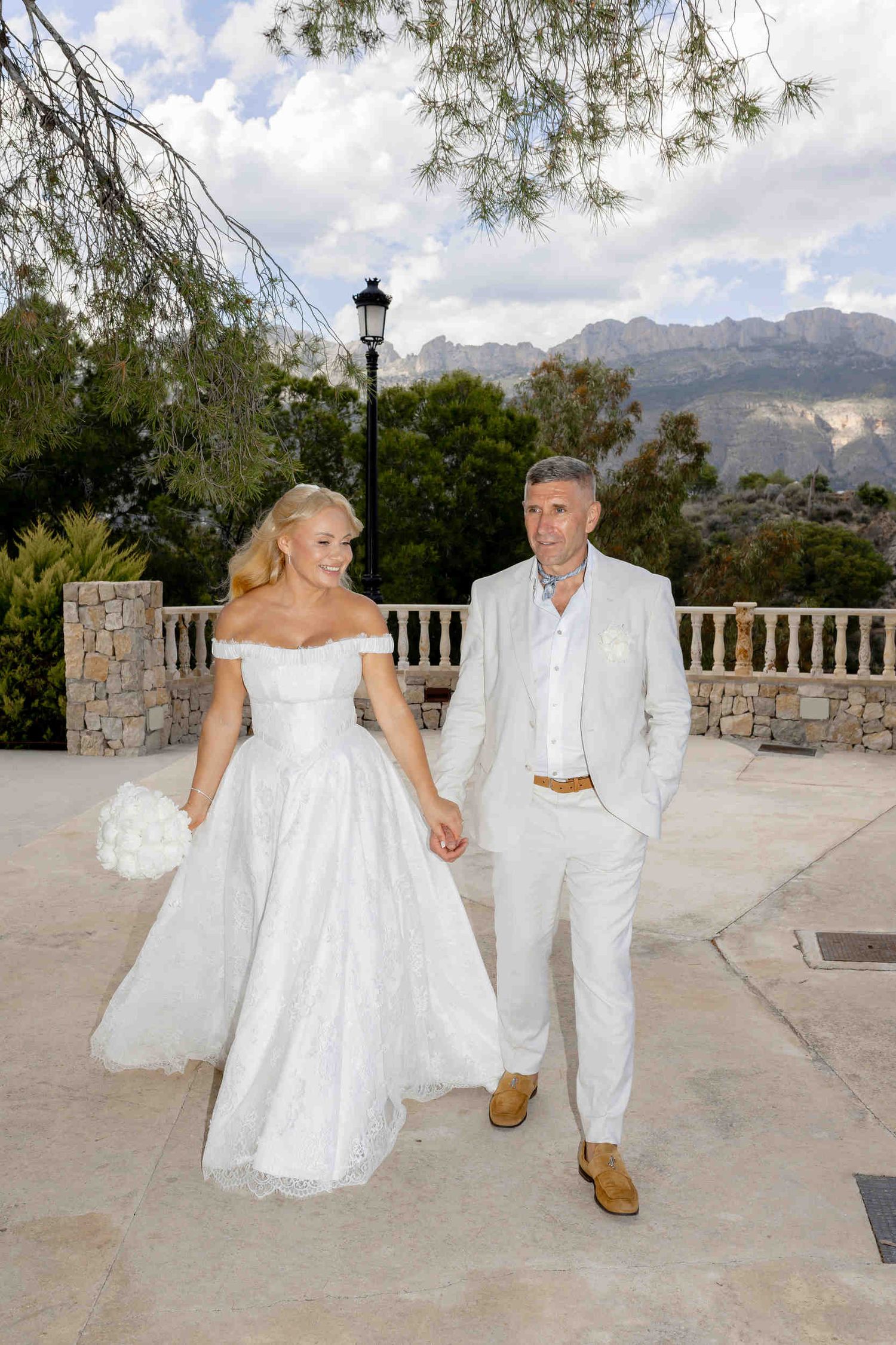 Happy bride in a lace dress and groom in a white suit walking together on a scenic terrace with mountain views in Altea.