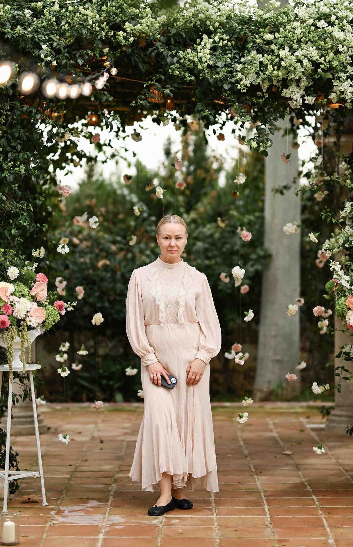 Wedding planner in Valencia Olga Prudko, stands under a garden floral archway in a beige dress holding her phone.