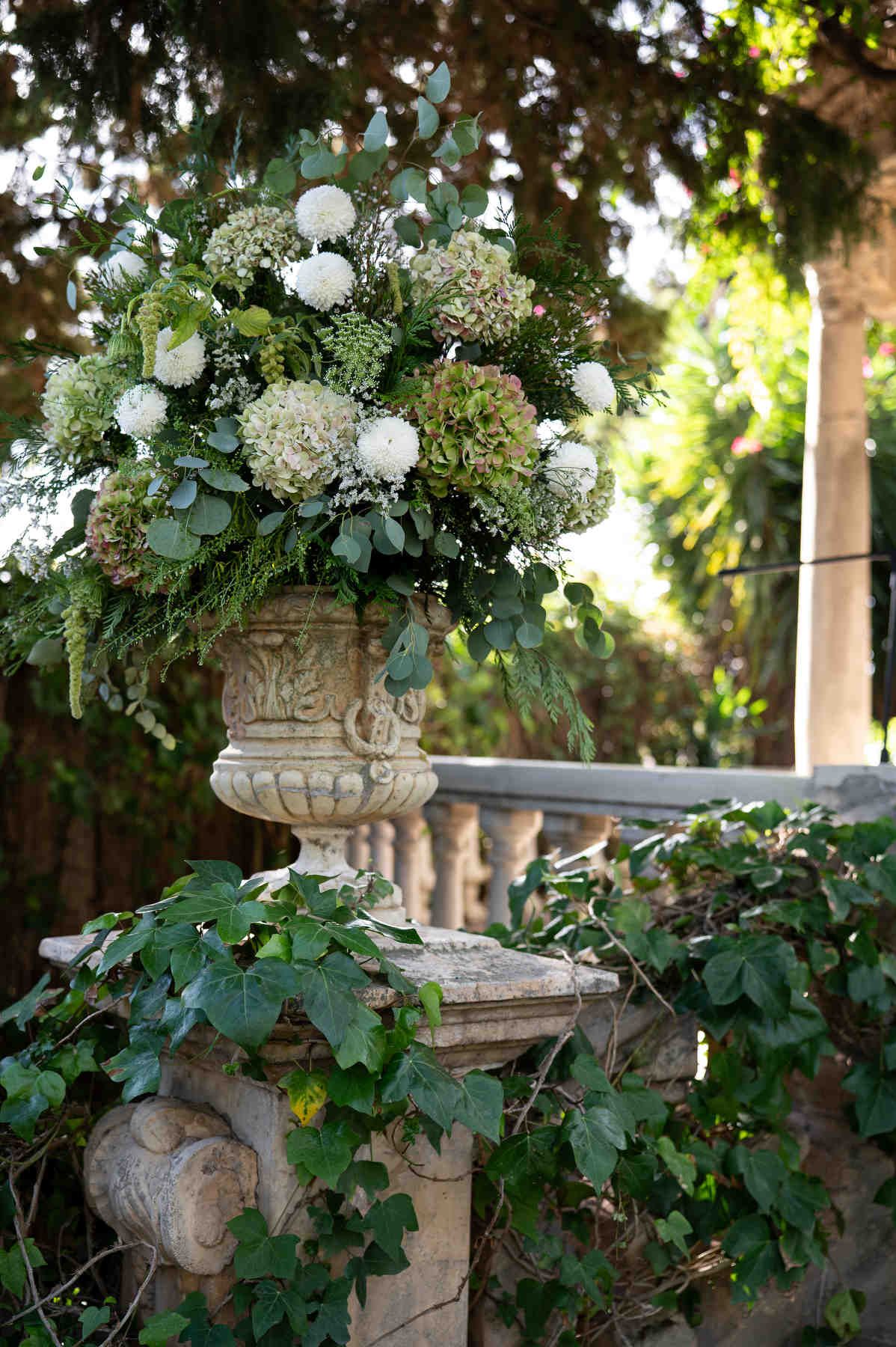 Elegant wedding floral arrangement in a vintage stone urn on an ivy-covered balustrade at the Casa Santonja estate.