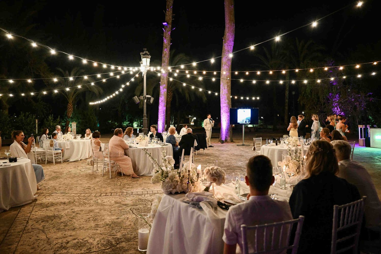 Host speaking to guests seated at illuminated round tables during a nighttime outdoor wedding reception under string lights in el Senyoret