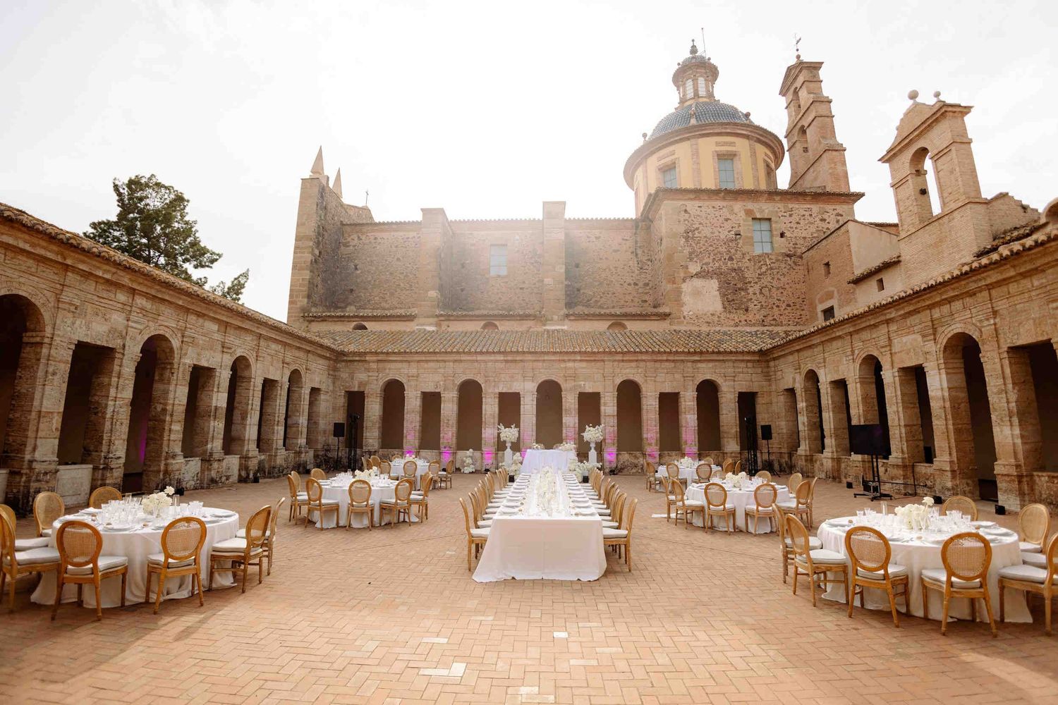 Outdoor wedding reception setup in the historic La Cartuja de Ara Christi courtyard, featuring round tables and pink uplighting.