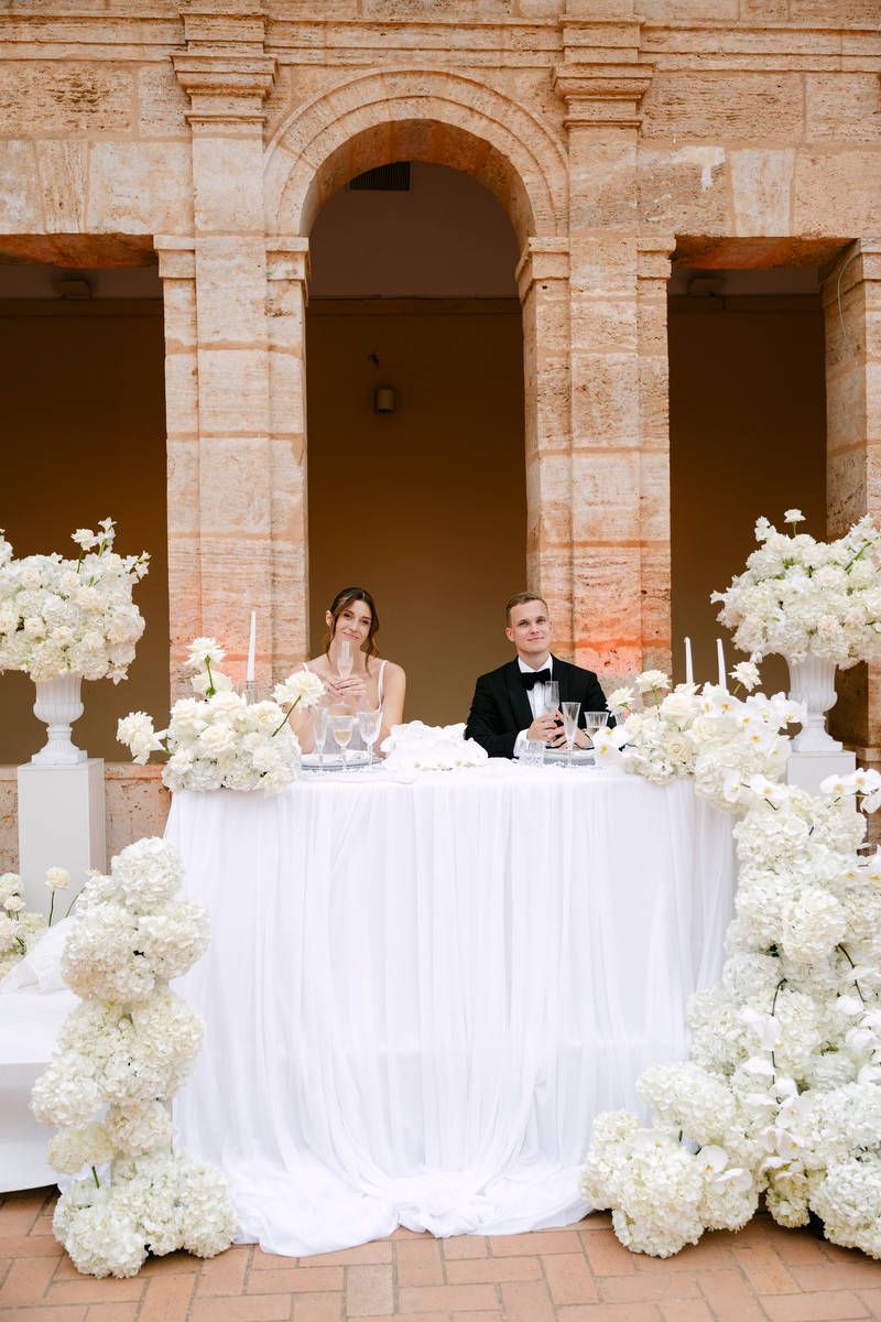 Bride and groom raising champagne glasses at a sweetheart table decorated with draped fabric and white florals in Valencia.