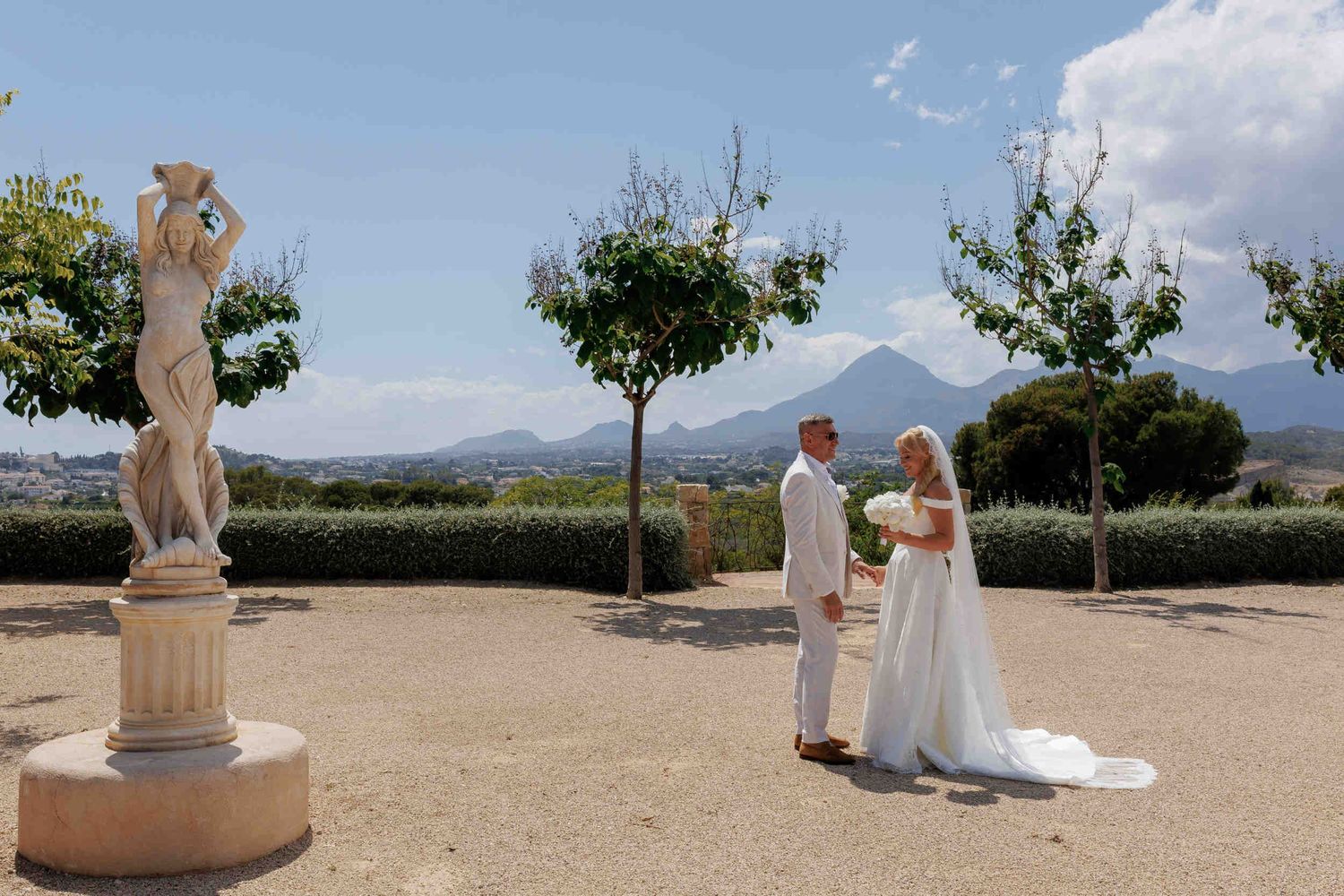 Groom in a white suit holding his bride's hand next to a stone statue with a scenic mountain view at a Costa Blanca finca.