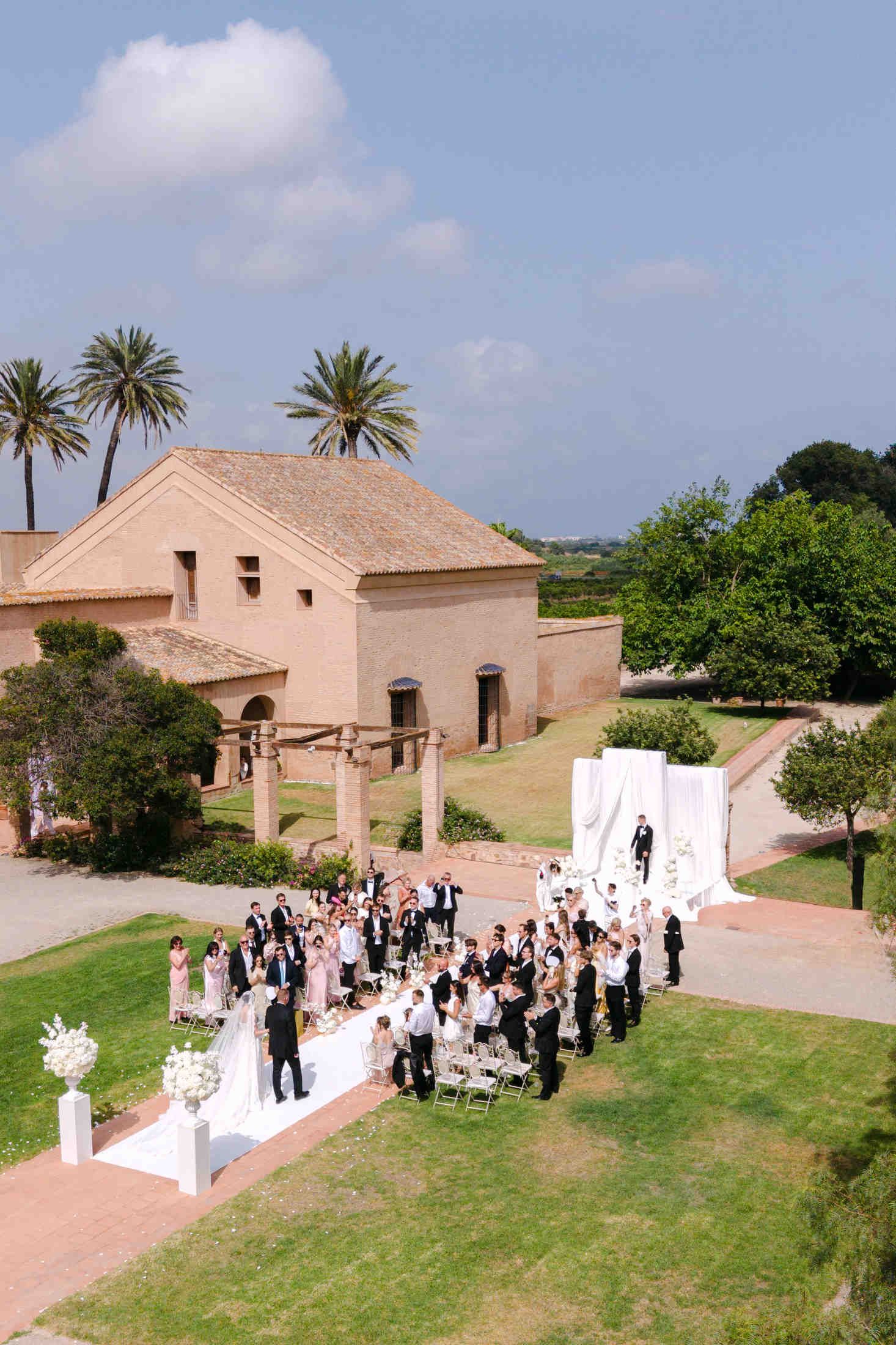 Destination wedding ceremony at a historic Valencia finca, showing guests gathered around a white aisle runner and palm trees.