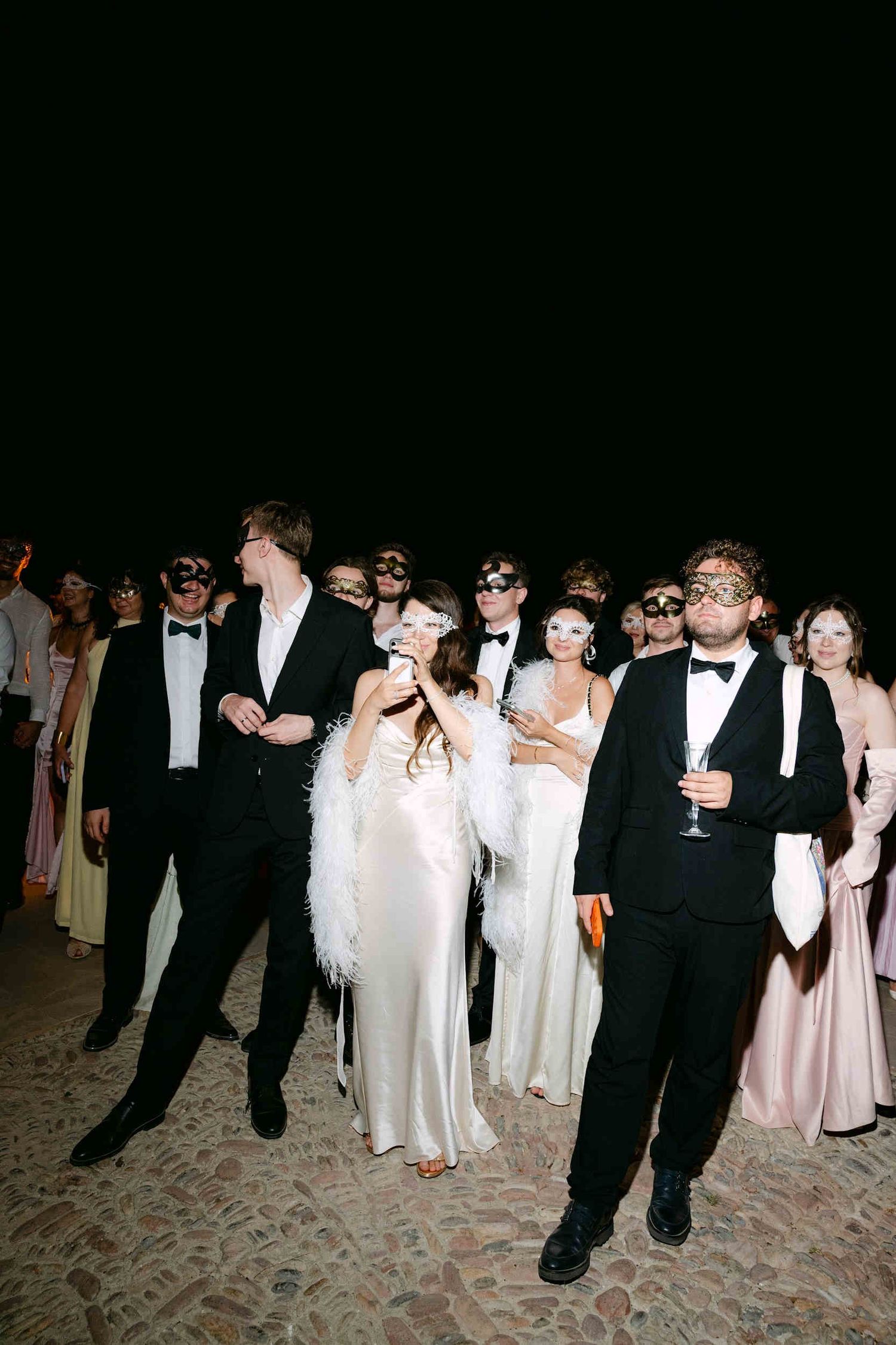 Wedding guests wearing elegant Venetian masquerade masks on a cobblestone courtyard during a night party at La Cartuja.