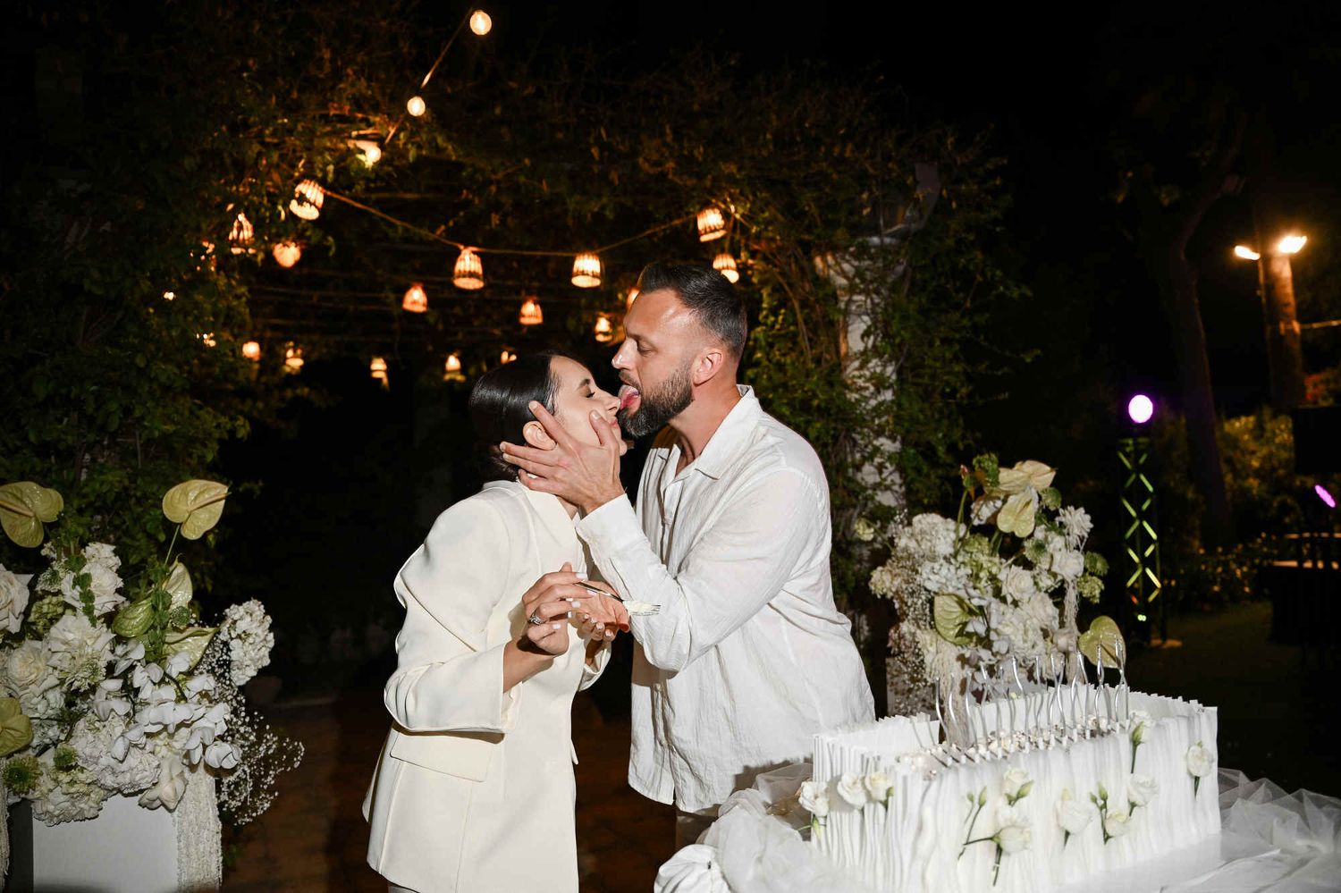 Groom playfully licking the bride's face while cutting their elegant white wedding cake at an outdoor evening reception in Valencia.