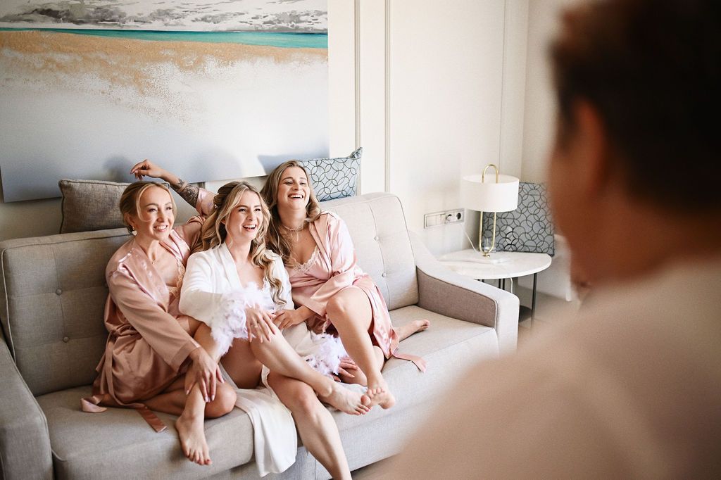 Bride and bridesmaids in silk robes laughing on a couch before their flawlessly planned destination wedding in Benidorm.