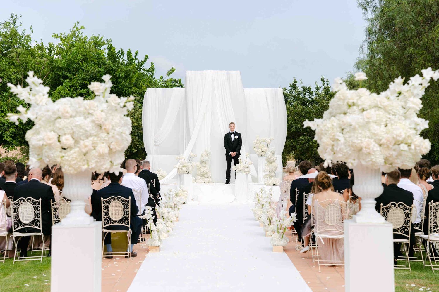 Groom in a black tuxedo waiting at a white draped floral altar during an outdoor ceremony at La Cartuja de Ara Christi.
