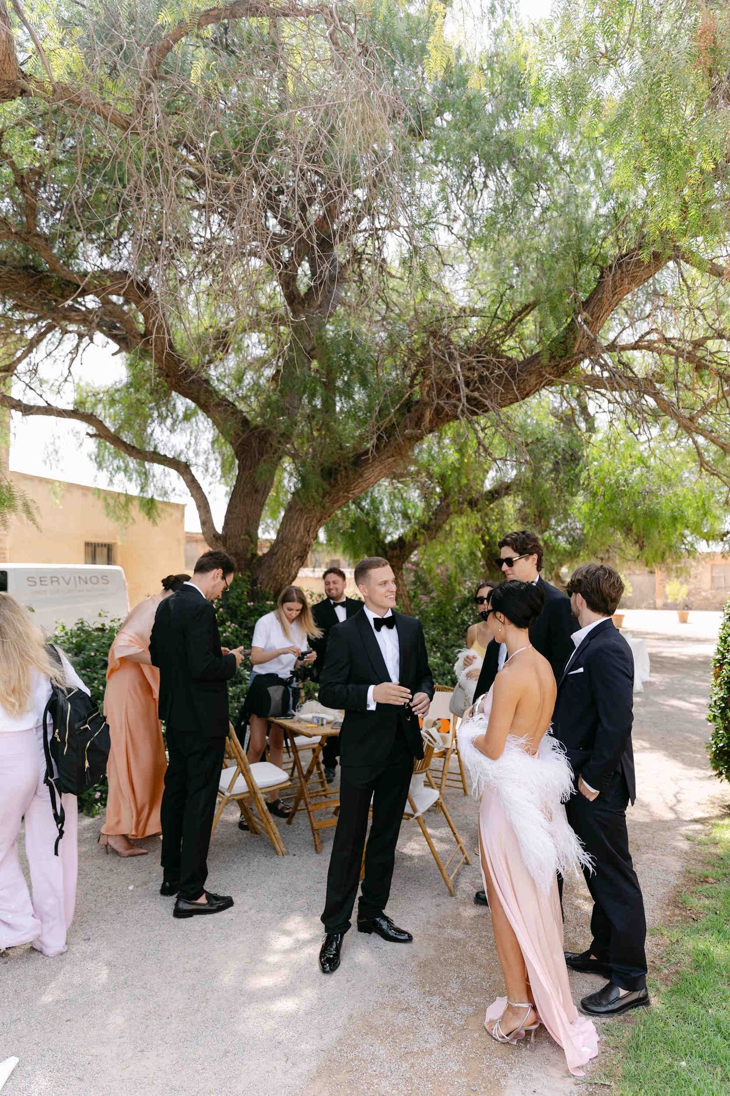Groom in a black tuxedo chatting with wedding guests under the shade of a large tree at La Cartuja de Ara Christi.