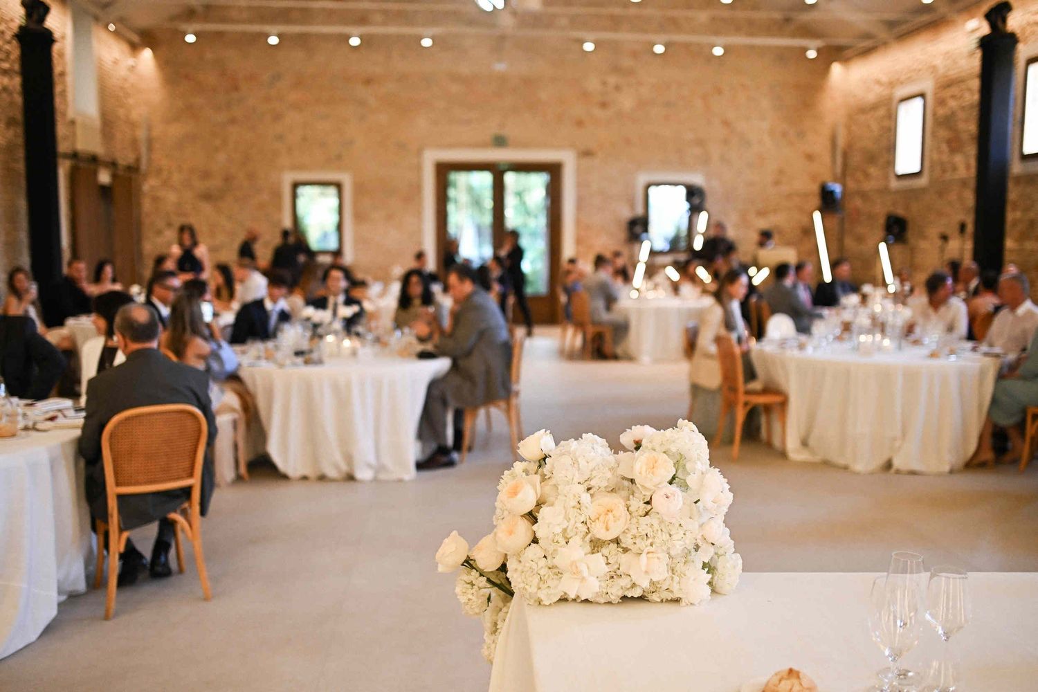 Lush white floral arrangement on a table with guests dining in the blurred background at an indoor Valencia wedding.