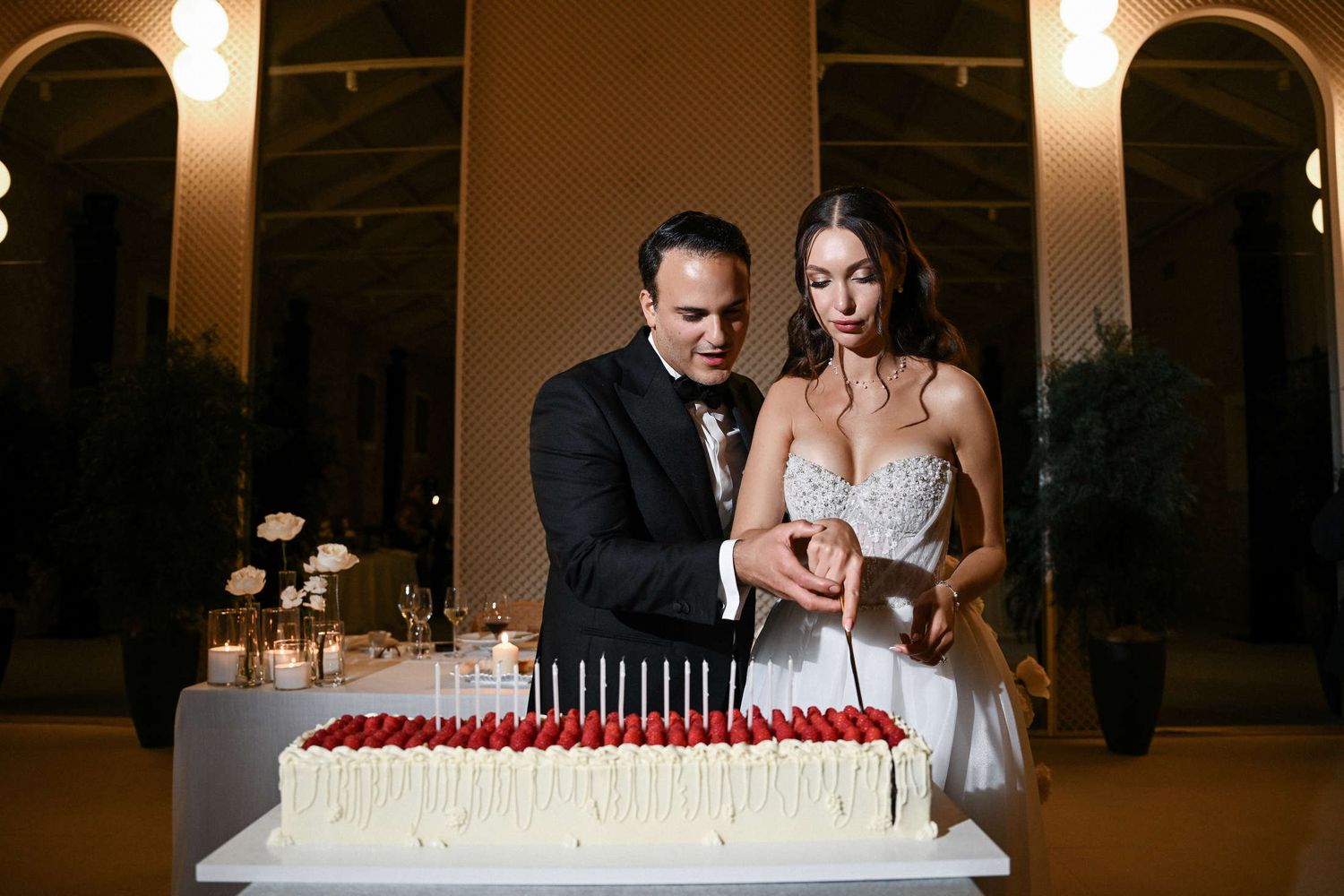 Bride and groom cutting a long rectangular wedding cake topped with red raspberries and candles at a Valencia event