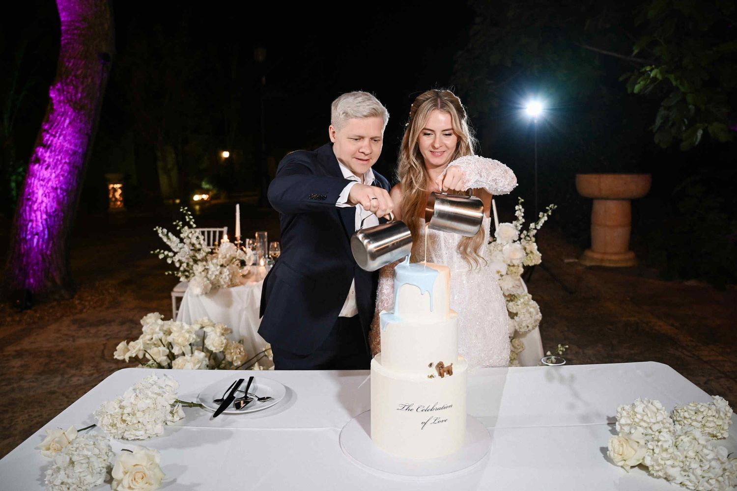 Bride and groom pouring colored glaze over their custom white wedding cake featuring a small dog figurine at their outdoor reception.