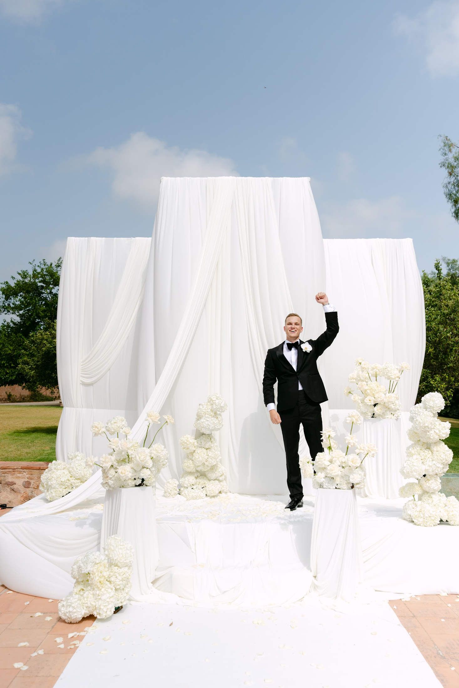 Groom celebrating on a white floral altar. Expert wedding planner Valencia design at an outdoor estate under blue skies.