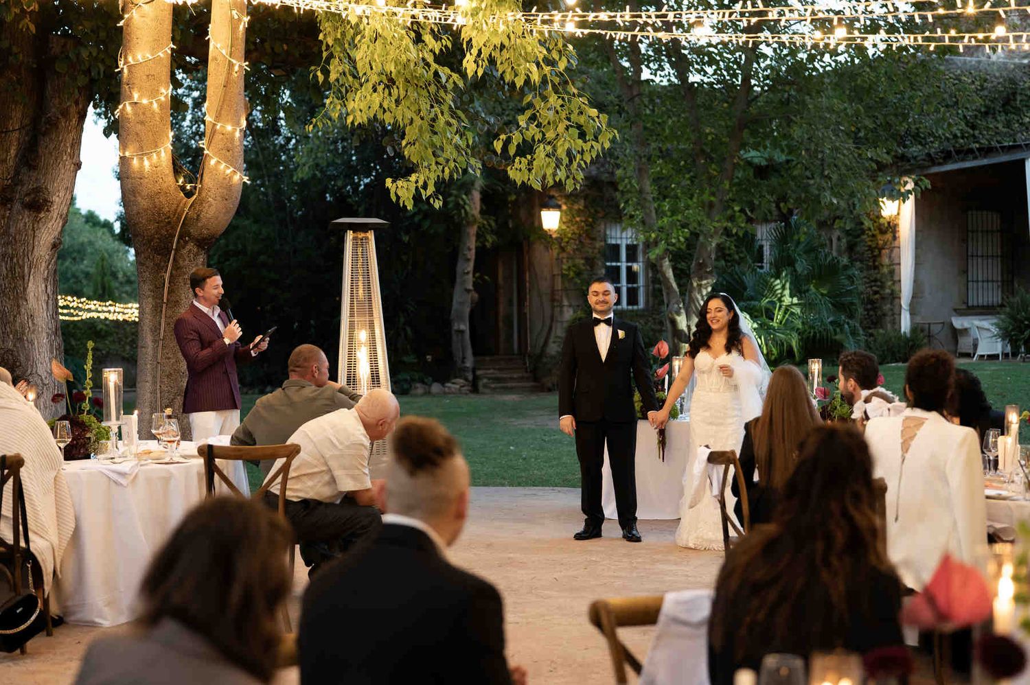 Wedding reception dinner outdoors under string lights with guests listening to speeches