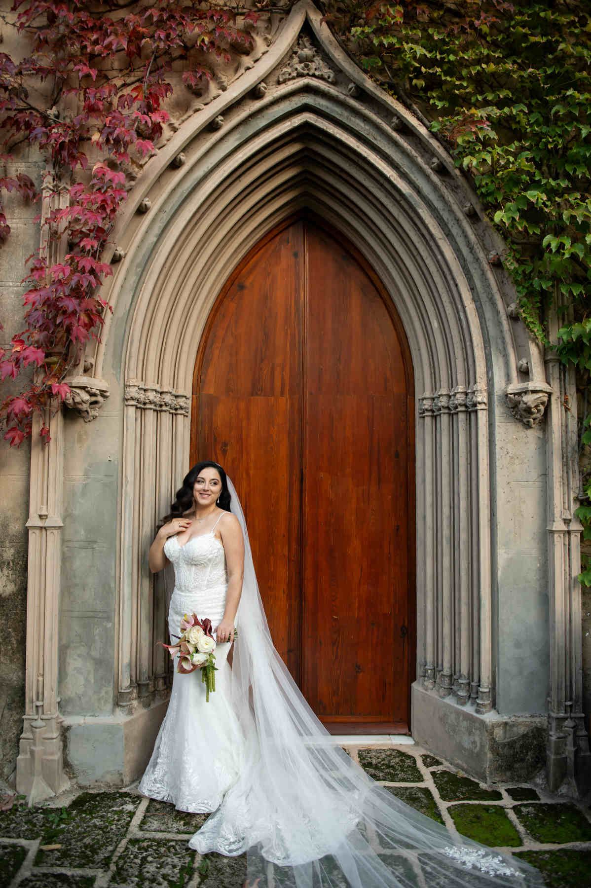 Elegant bride in a mermaid wedding dress with a long veil standing by the wooden doors of Casa Santonja.