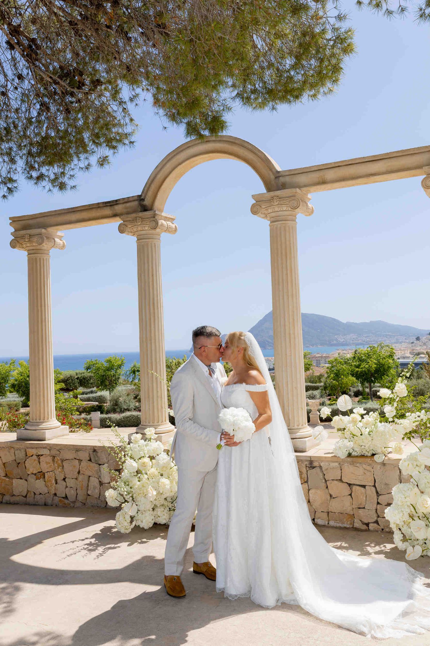 Bride and groom kissing under a stone arch with sea view during their luxury wedding ceremony in Altea.