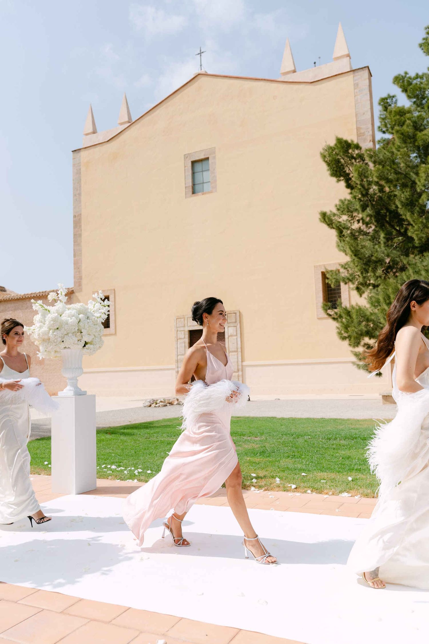 Smiling bridesmaids wearing pastel silk dresses and feather boas walking down a white ceremony aisle at La Cartuja.