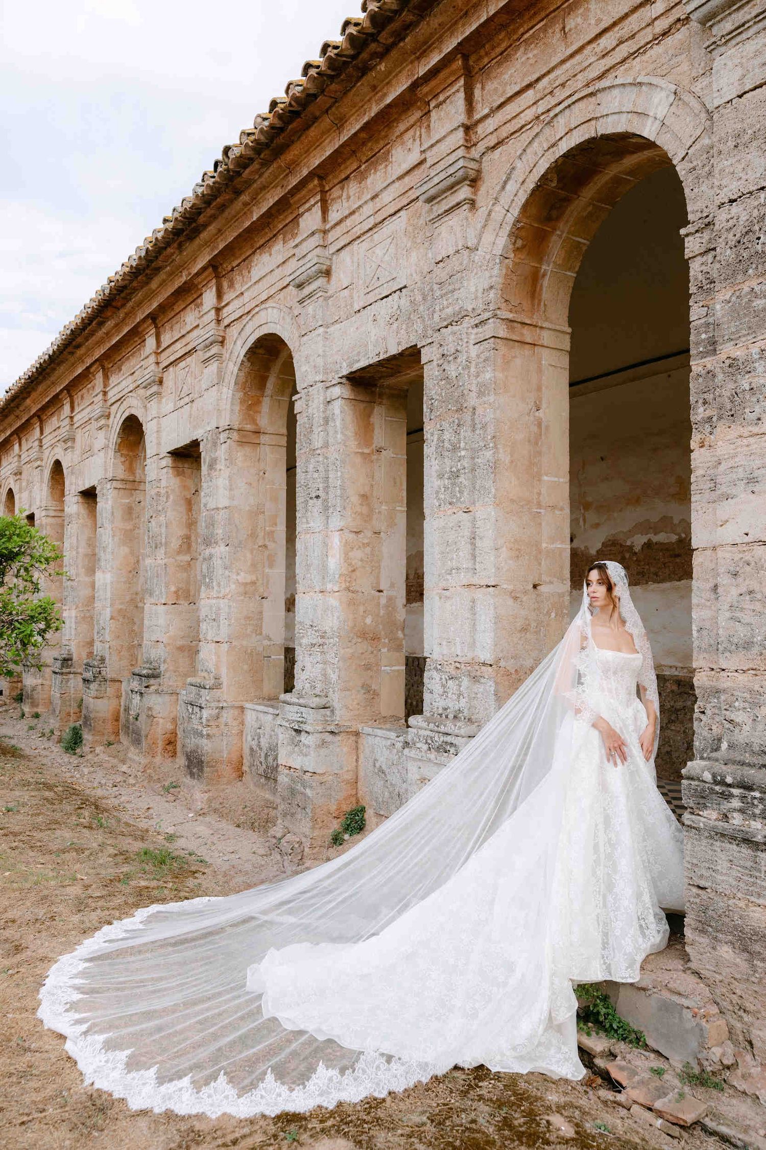 Bride poses alone inside an ancient stone cloister archway, showcasing her long lace veil and extended wedding dress train.
