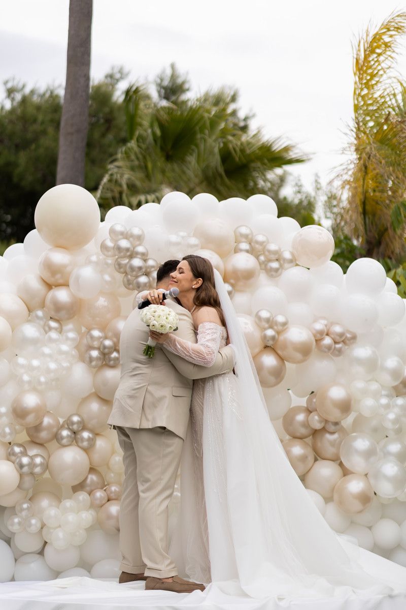 Bride holding a microphone hugs her groom near a white balloon arch at an event styled by an Alicante wedding planner.