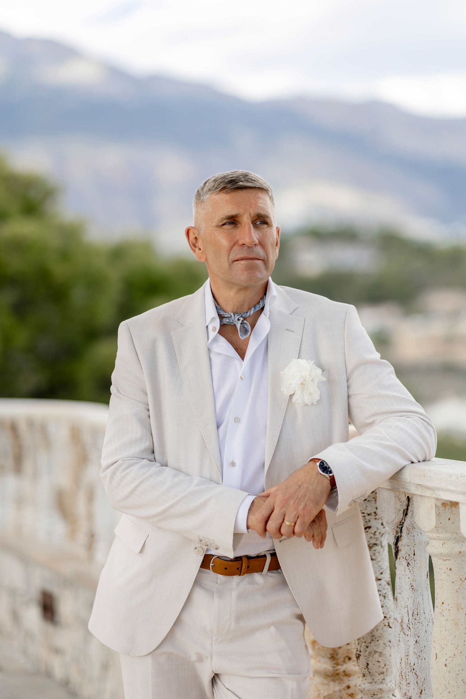 Groom in an elegant white suit posing on a stone balcony overlooking the Spanish landscape at Finca Marques de Montemolar in Altea.